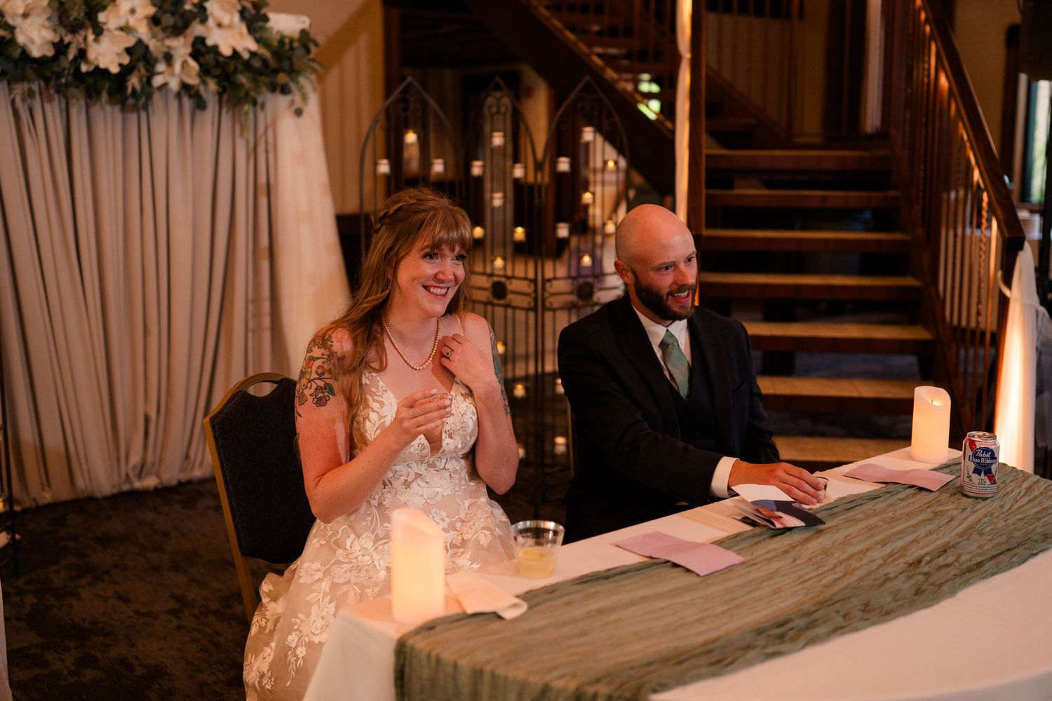 Bride laughing with groom at sweetheart table during romantic candlelit wedding reception.
