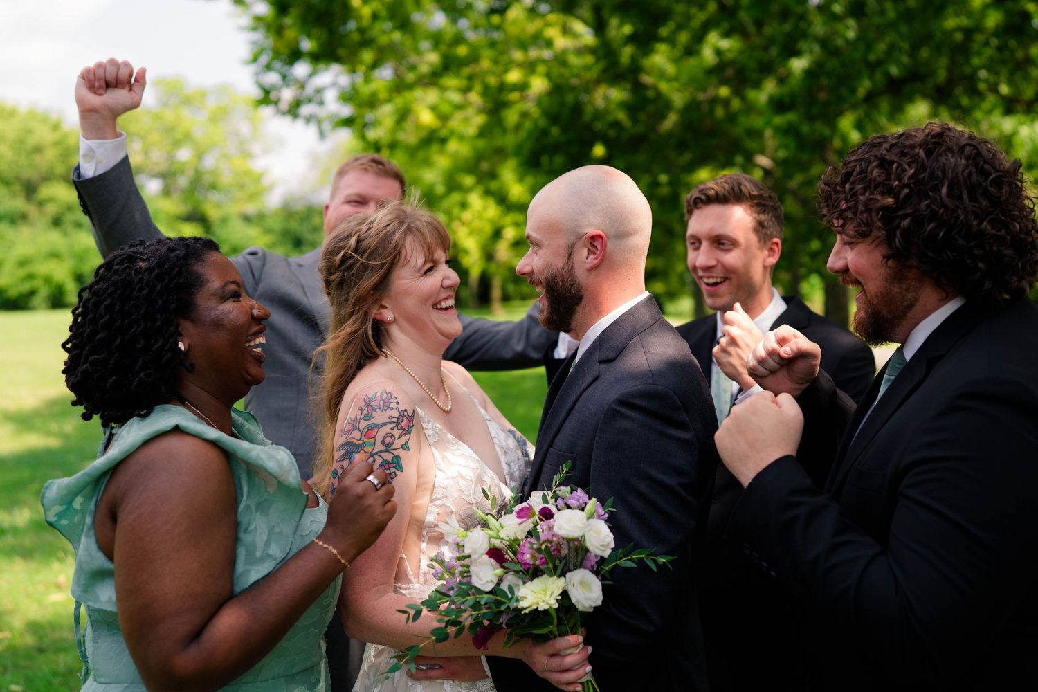 Joyful wedding party celebrates with bride and groom outdoors, guests cheering and laughing in green park setting.