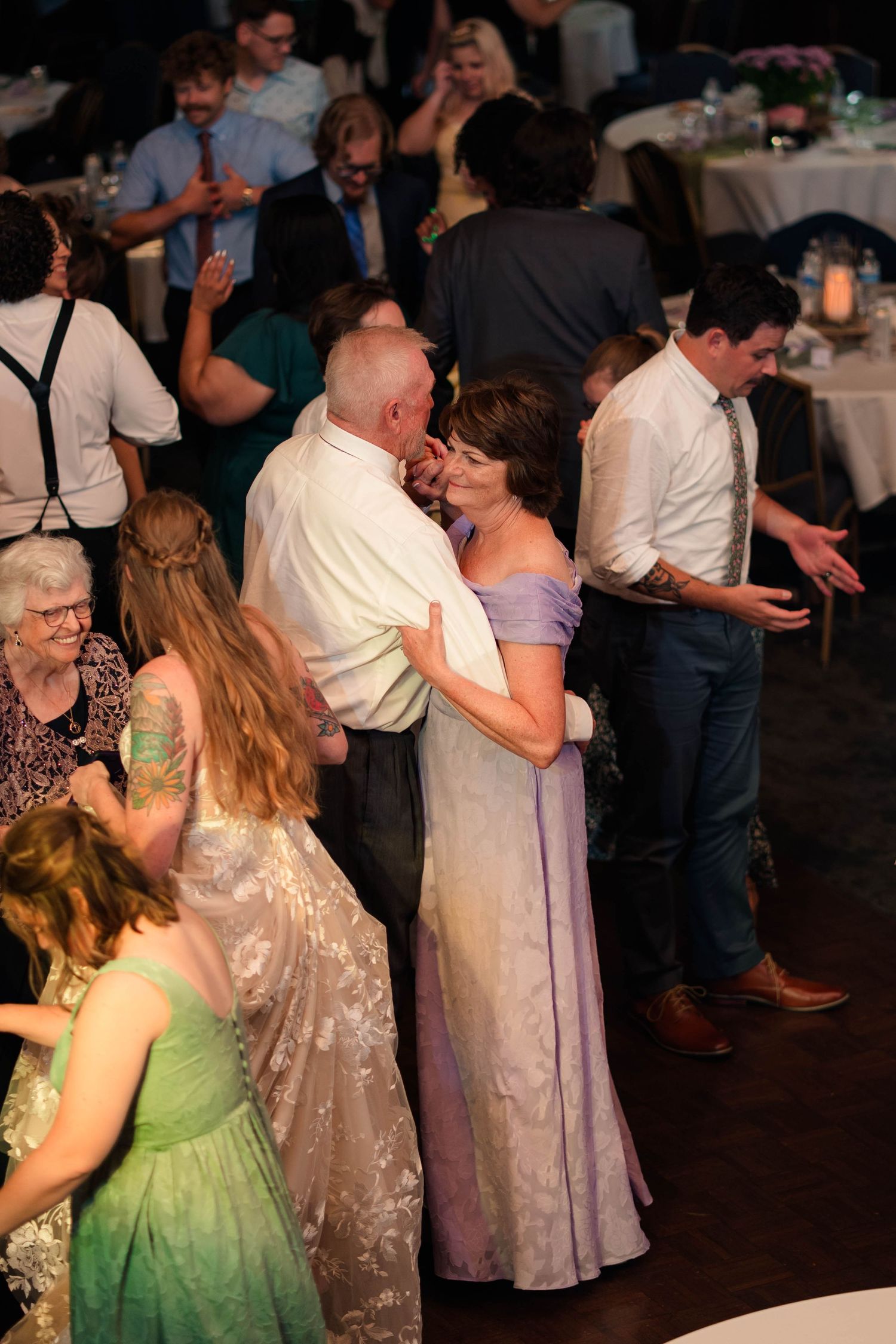 Couple in formal attire dance closely at crowded wedding reception with guests dancing around them.