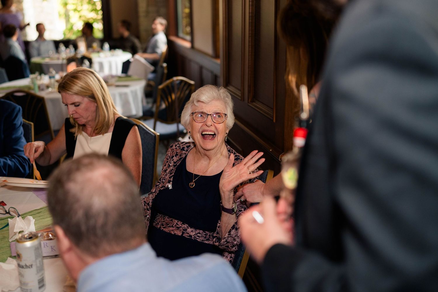 Elderly woman laughing and waving at wedding reception dinner table surrounded by guests.