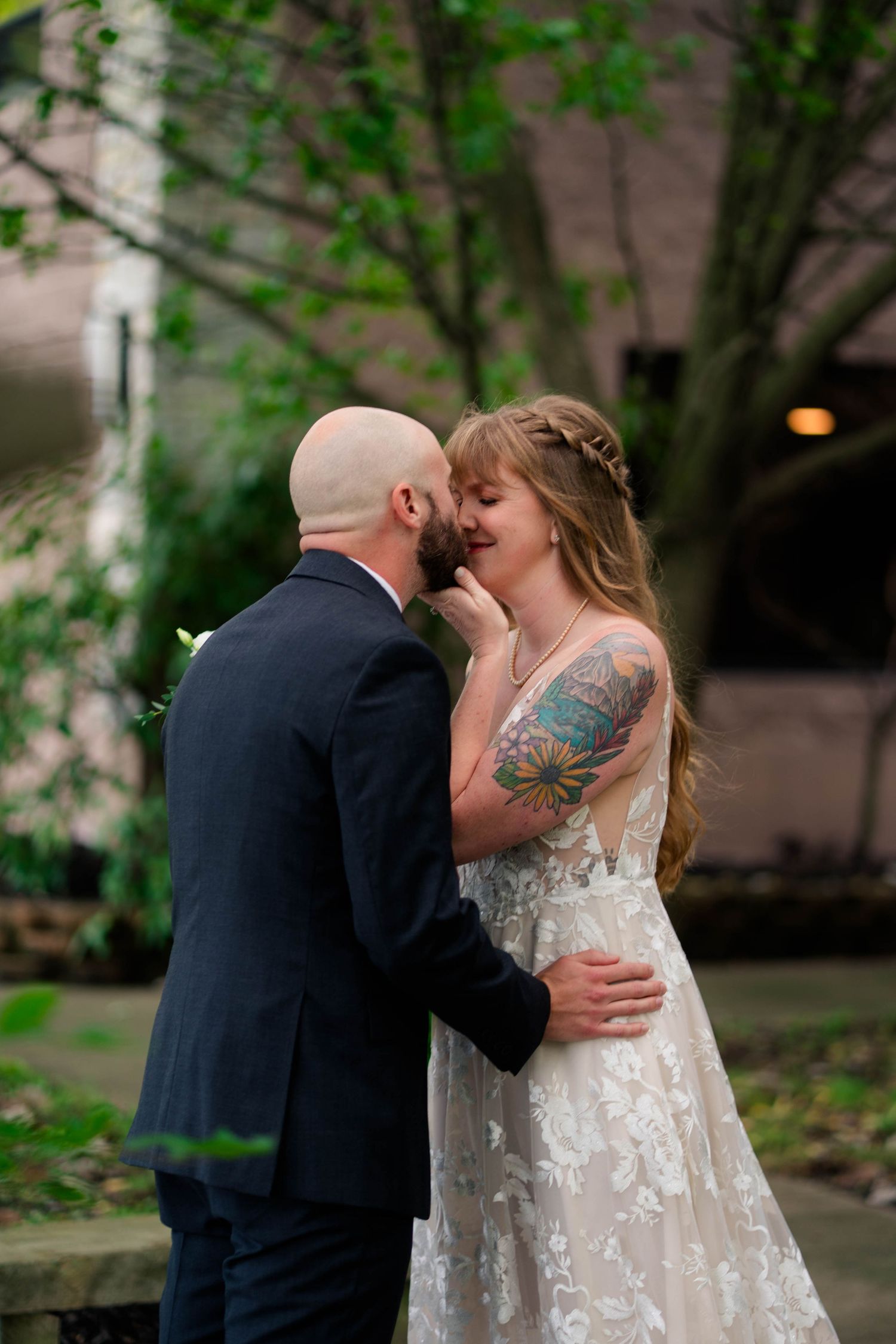 Tattooed bride and bald groom share intimate moment outdoors surrounded by lush green trees.