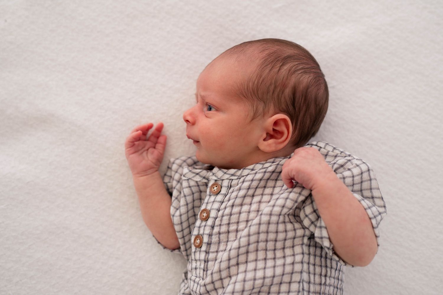A peaceful newborn baby in a plaid onesie lies on a white textured blanket with tiny hands raised.