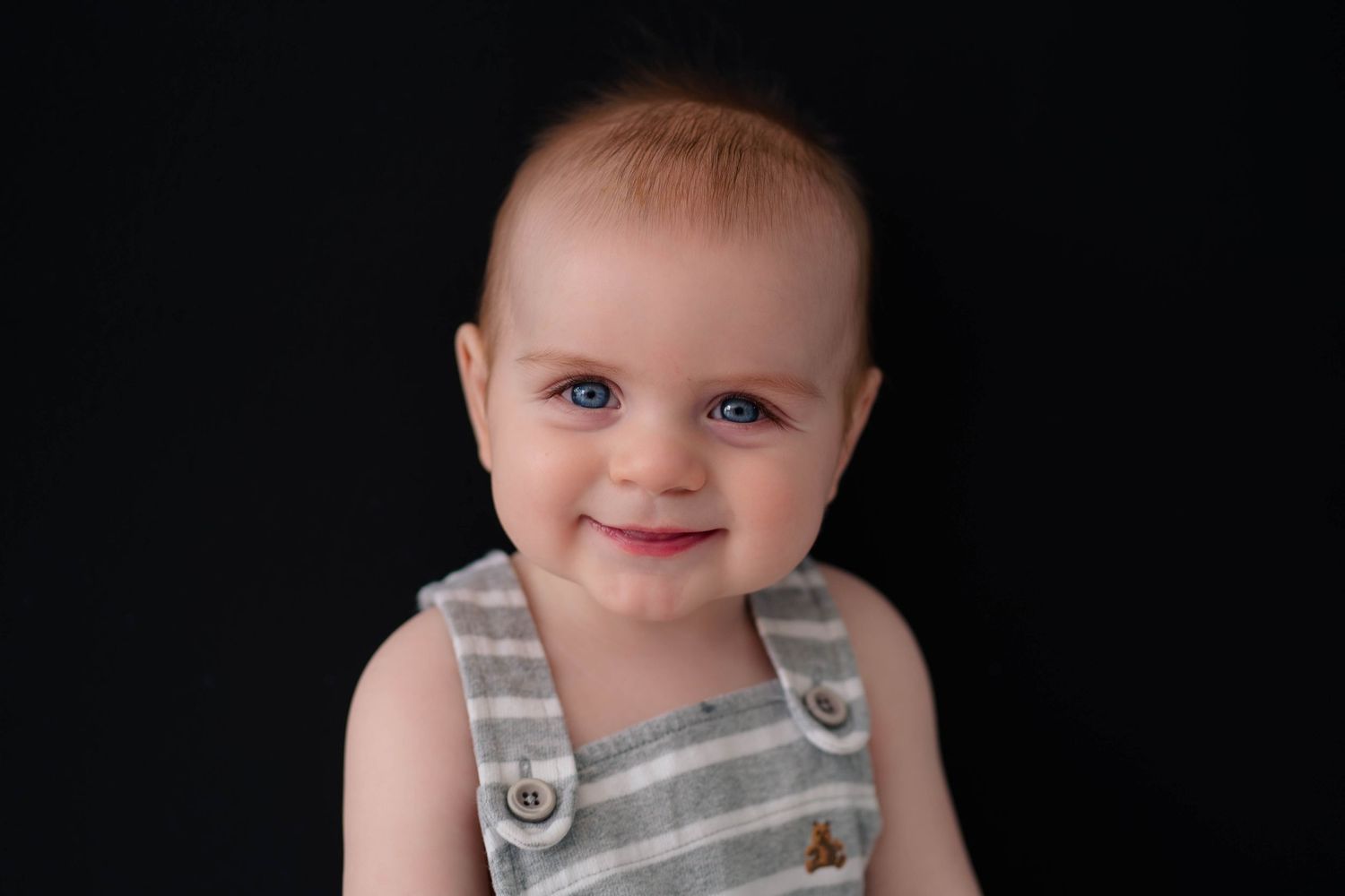 Adorable blue-eyed baby smiling in a gray striped romper against a dark background.