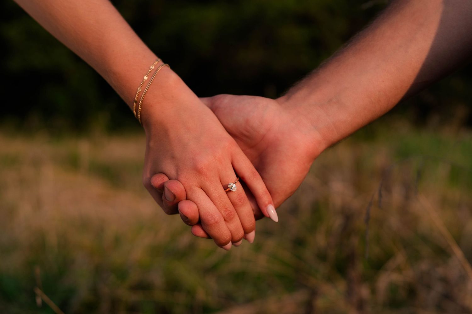 Two people holding hands outdoors, one wearing a diamond engagement ring and gold bracelet, with a blurred green field background.