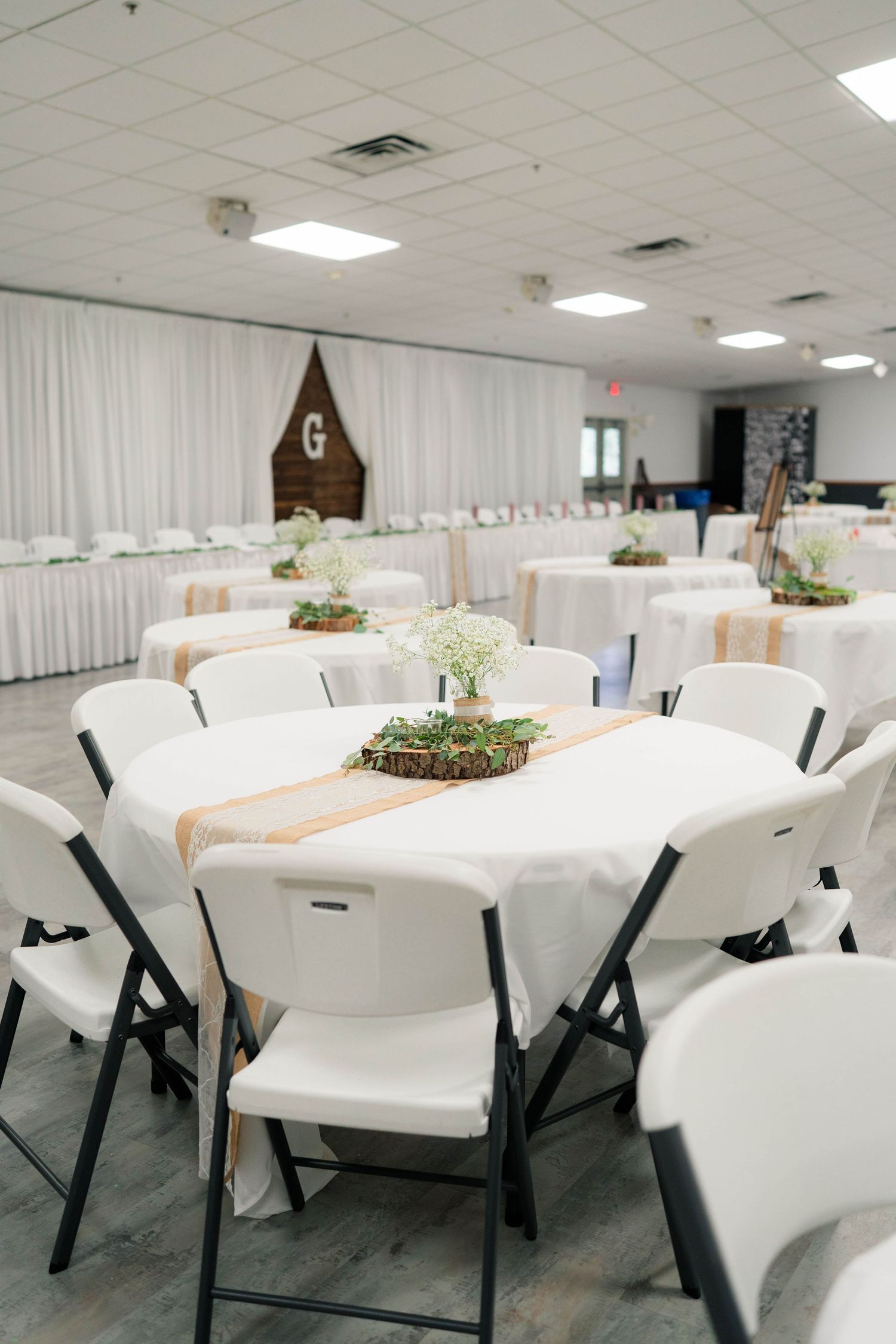 Elegant wedding reception hall with round tables draped in white linens, burlap runners, and floral centerpieces.
