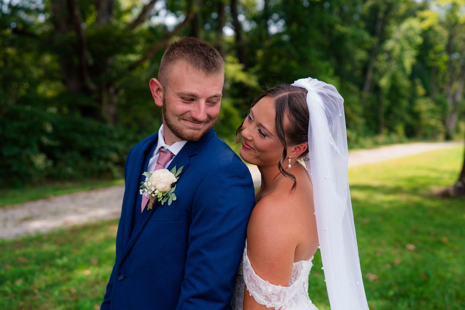 Bride in lace gown and groom in navy suit share a joyful outdoor moment surrounded by lush green trees.