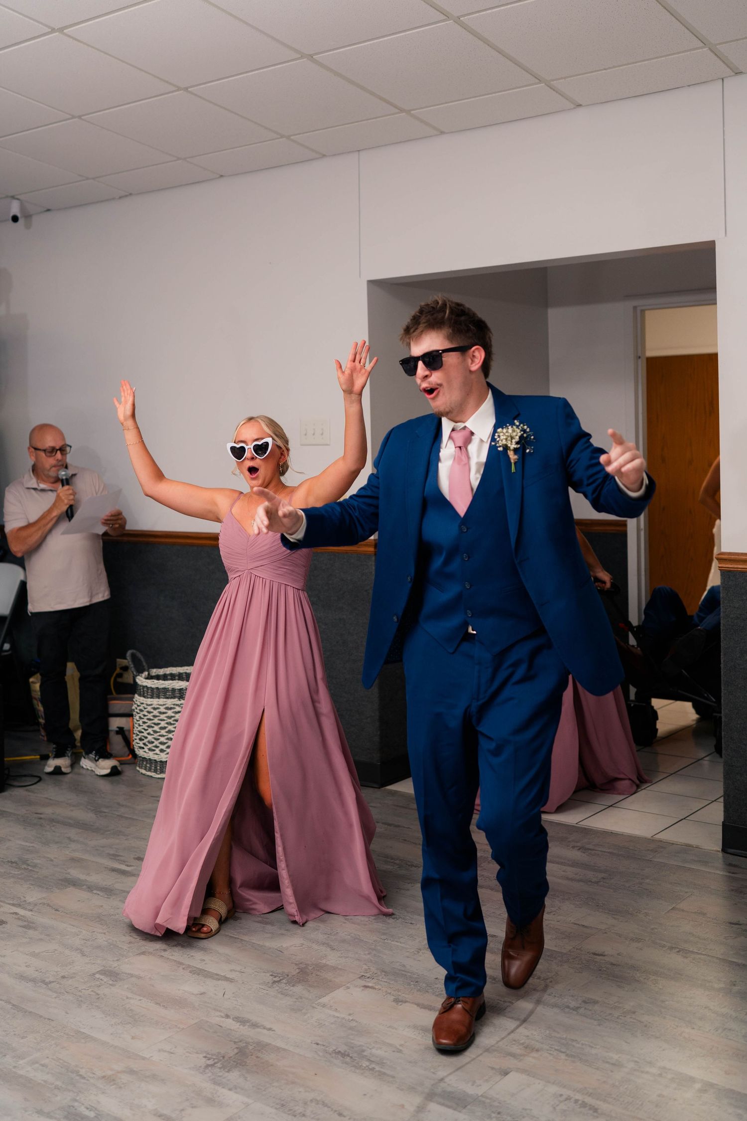 Groomsman in navy suit and bridesmaid in pink dress wearing sunglasses dancing energetically at a wedding reception.
