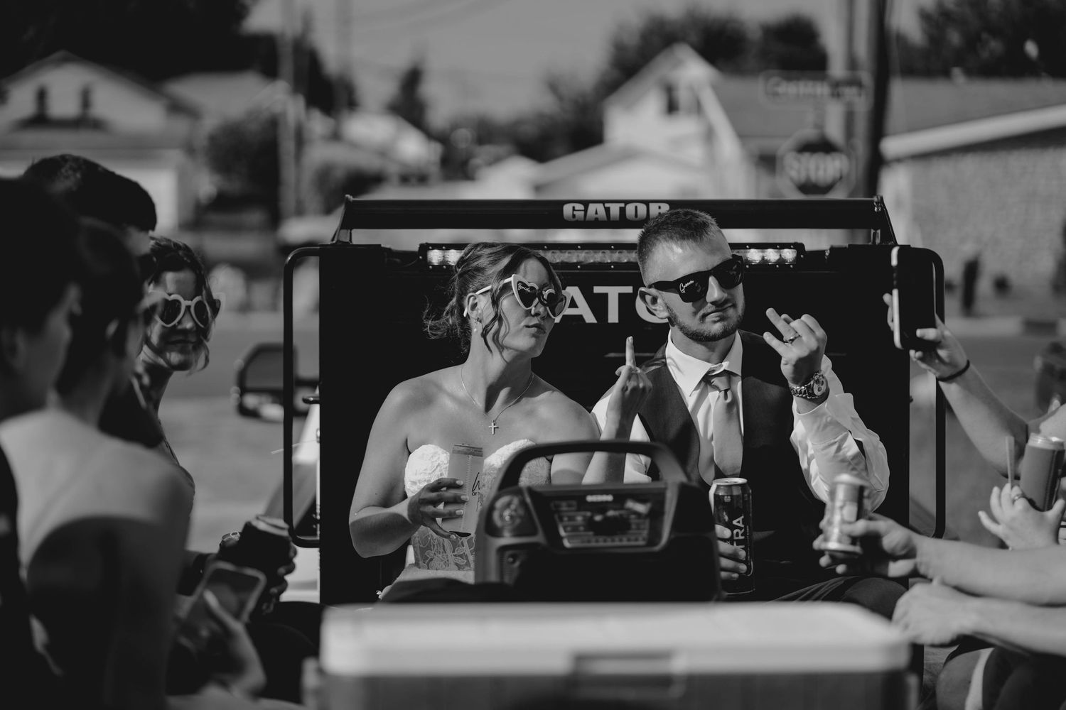 Black and white photo of bride and groom wearing sunglasses on a tractor with wedding guests.