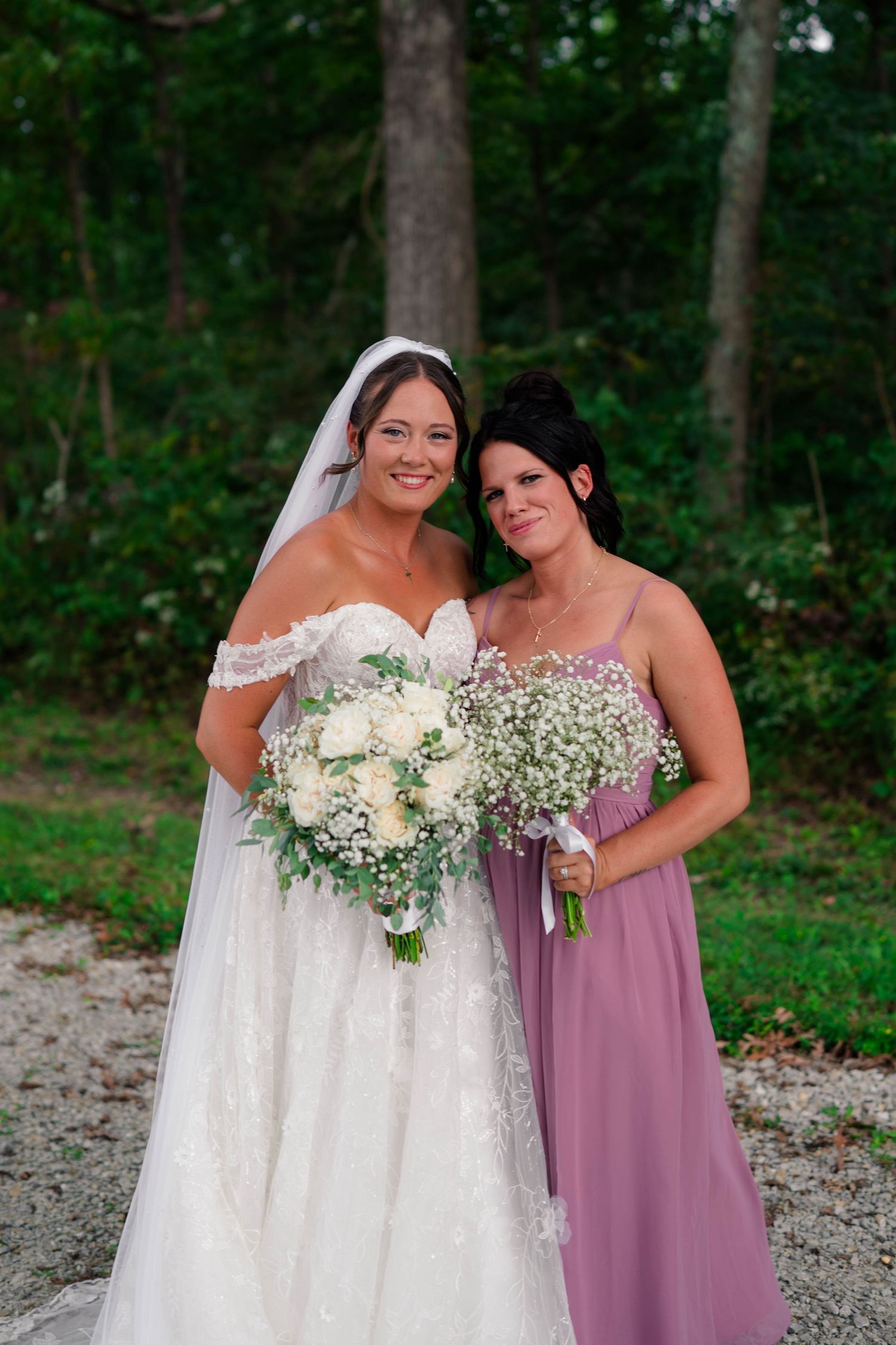 Smiling bride in off-shoulder lace gown poses with bridesmaid in mauve dress holding floral bouquets outdoors.