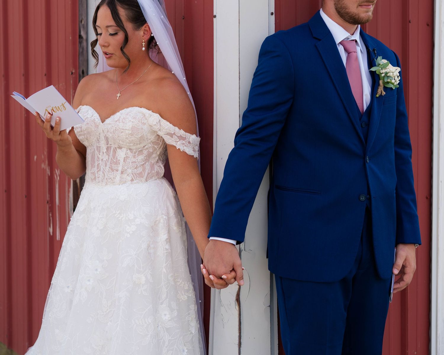 Bride reading vows while groom listens during romantic first look beside a red barn wall.