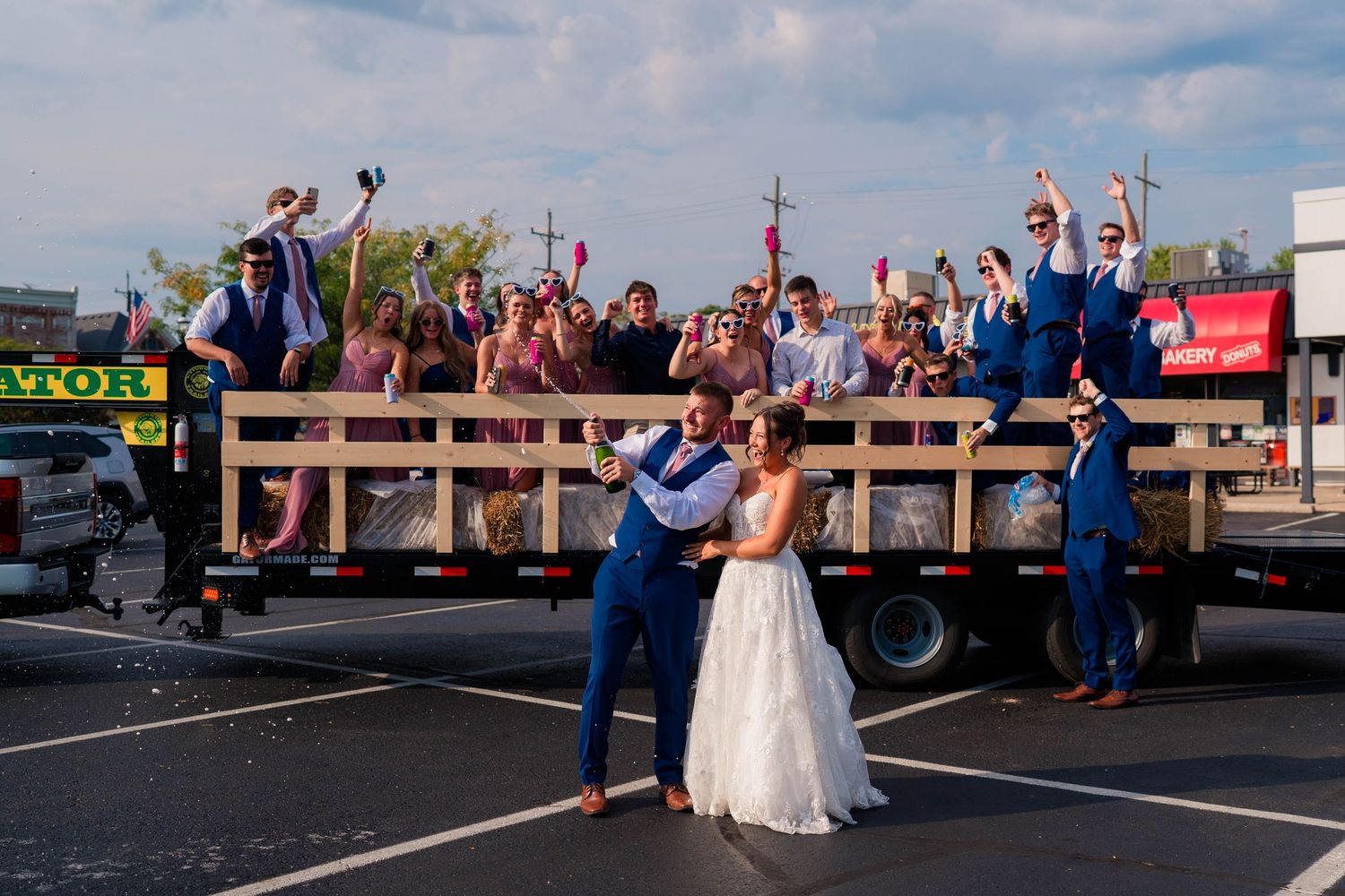 Bride and groom posing in front of flatbed truck filled with cheering wedding party.