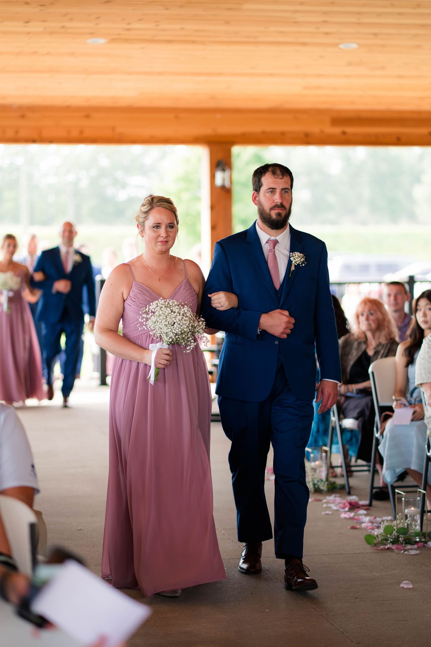 Bridesmaid in mauve gown walks down aisle with groomsman in navy suit during wedding ceremony.