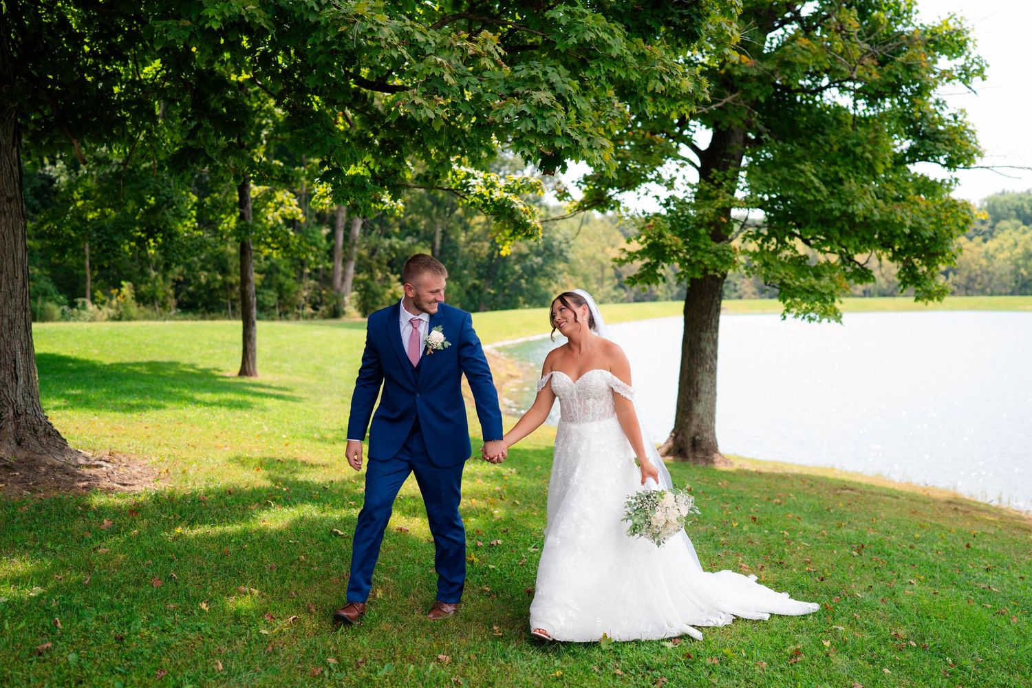 Bride and groom walking hand in hand on green lawn near a lake on their wedding day.