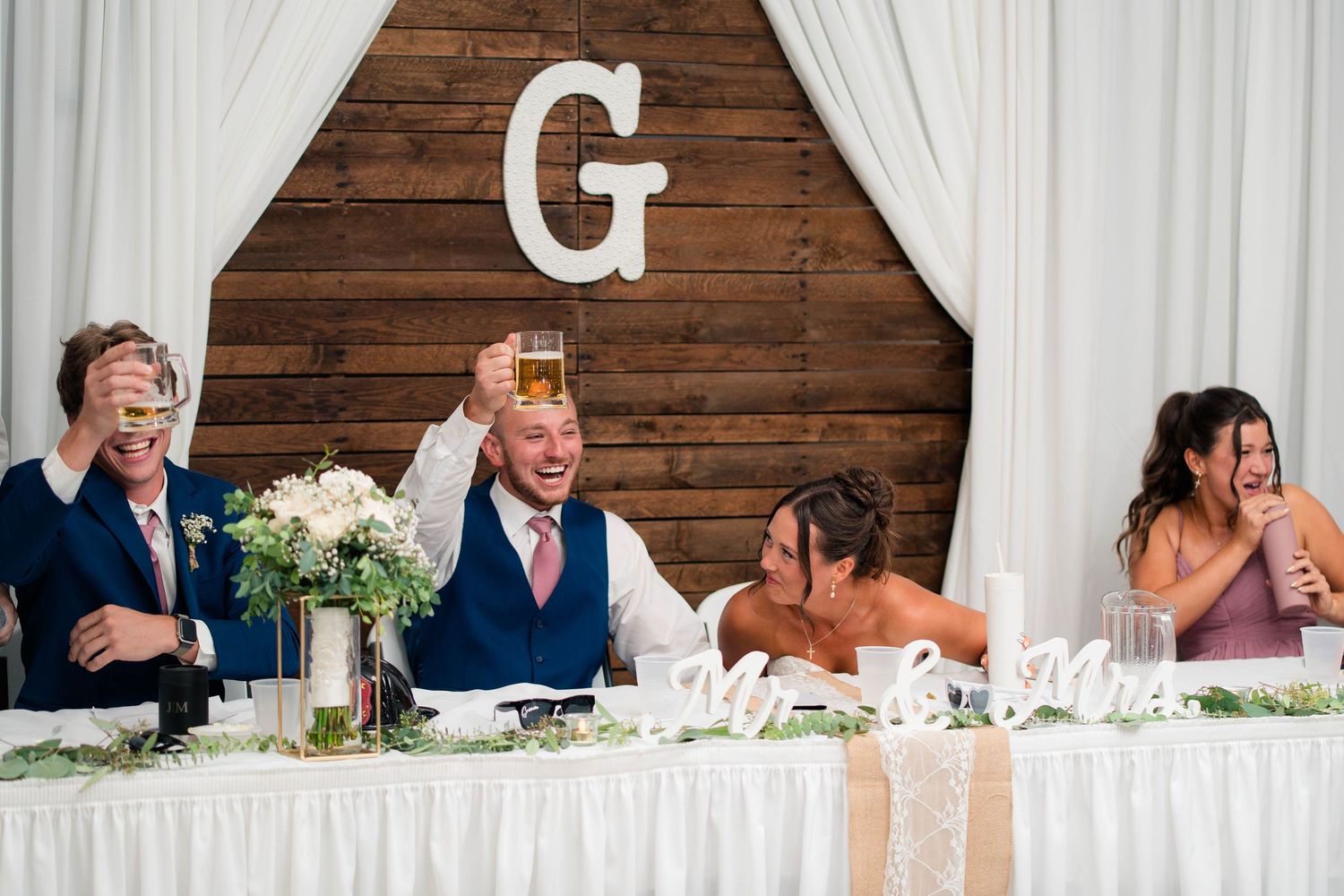 Wedding party raises glasses in a toast at the head table decorated with white florals and a large letter G backdrop.