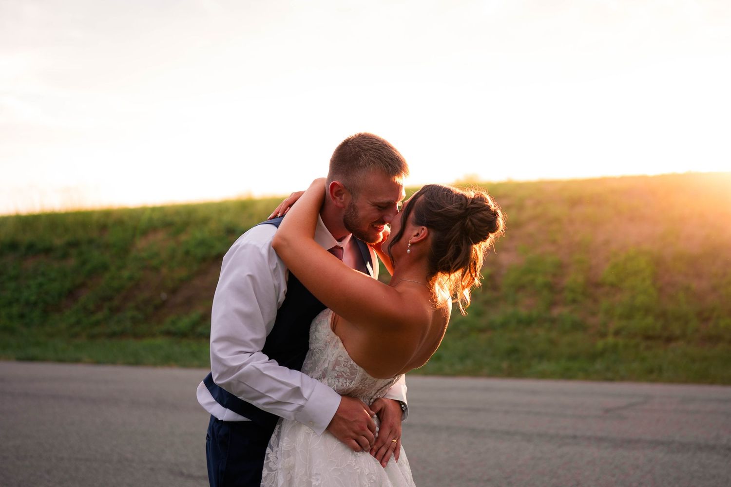 Bride and groom sharing a tender kiss outdoors at golden hour, bathed in warm sunset light.