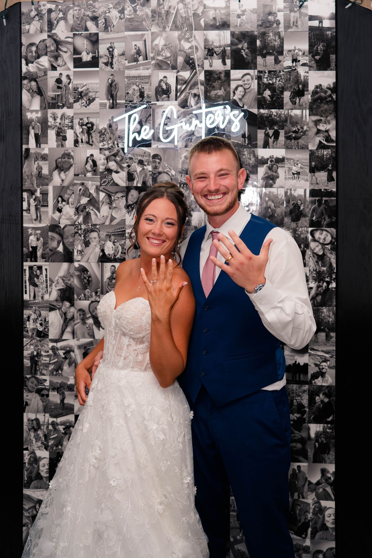 Newlyweds show off wedding rings in front of black and white photo collage backdrop with neon sign reading The Gunters.