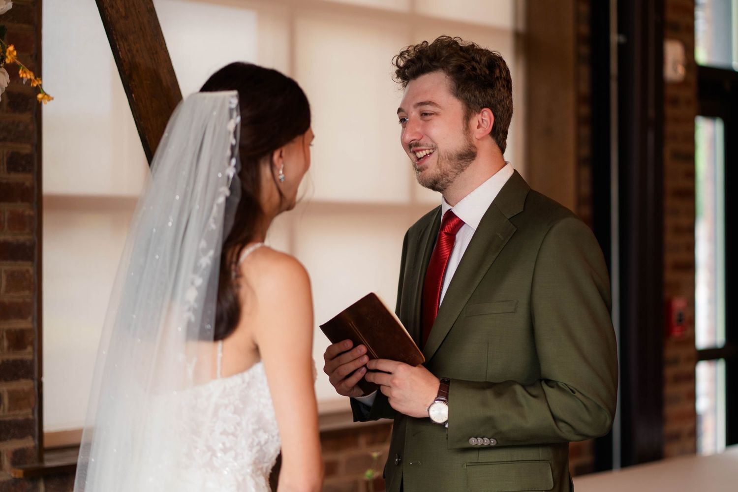 Smiling groom in olive suit reads vows to bride during intimate indoor wedding ceremony.
