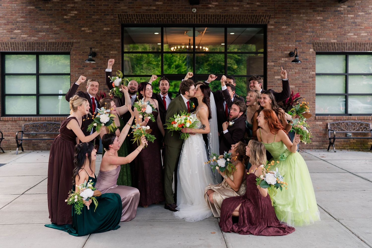 Bridesmaids in mismatched green and plum dresses celebrate joyfully with bride and groom outside brick venue.