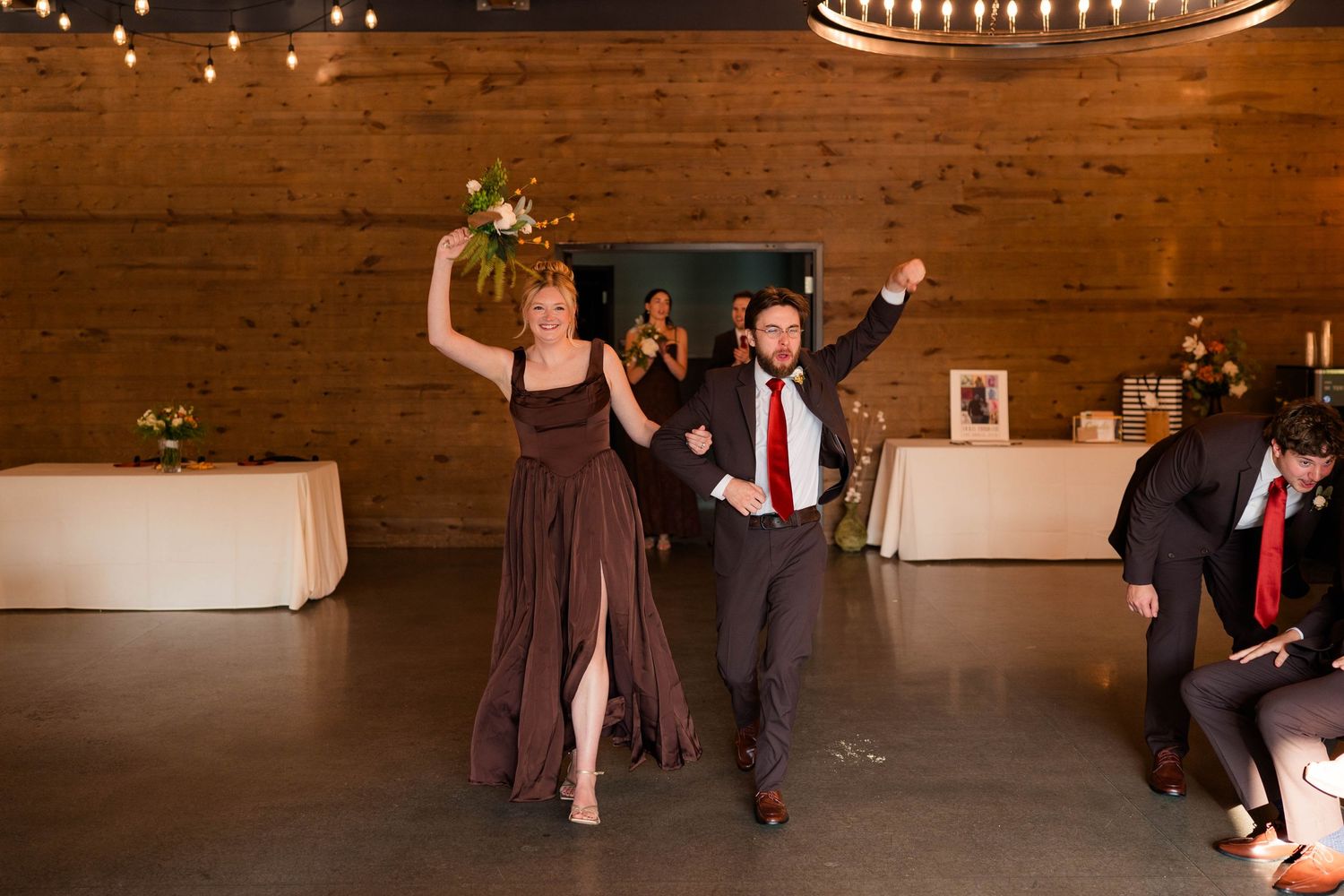 Bridesmaid in brown gown and groomsman make grand entrance into rustic wedding reception venue with string lights.