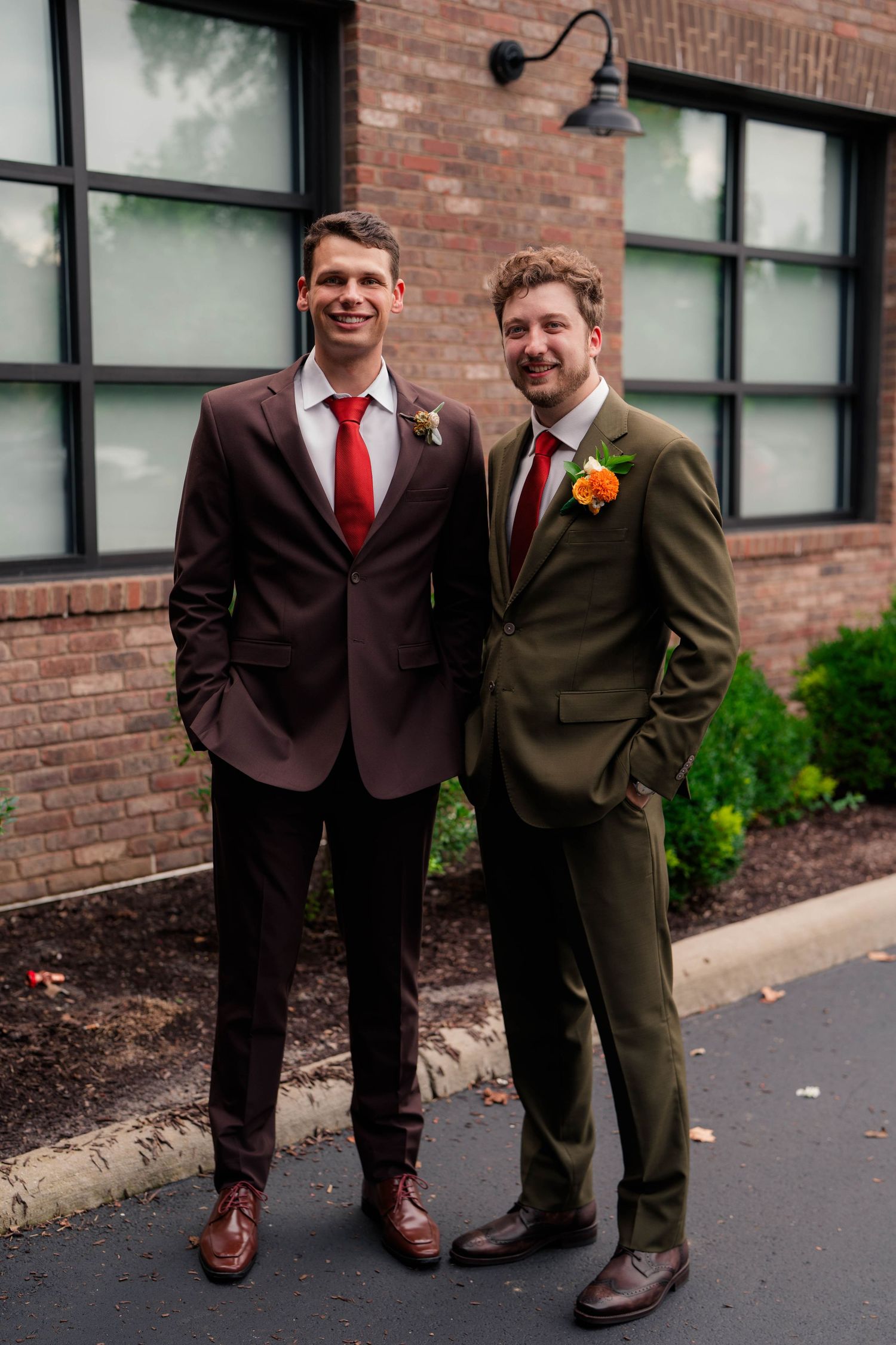 Two groomsmen pose outside a brick building, wearing olive and burgundy suits with floral boutonnieres.