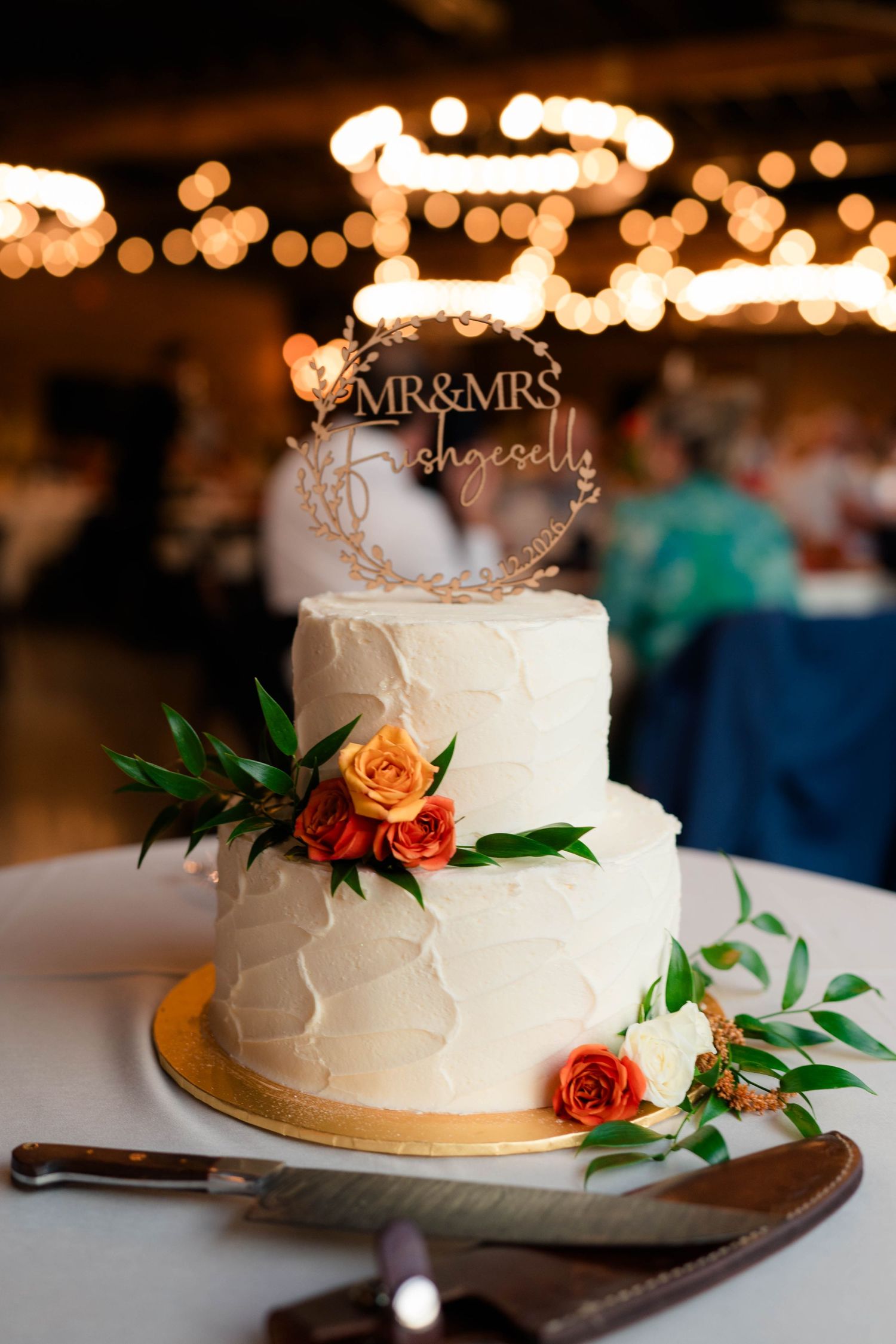Two-tier white wedding cake with orange roses and Mr and Mrs cake topper under warm string lights.