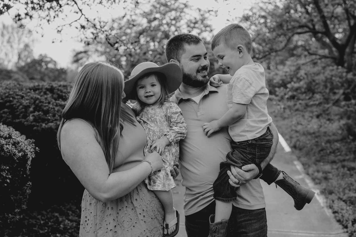 Black and white family portrait of parents holding two young children outdoors among lush greenery.