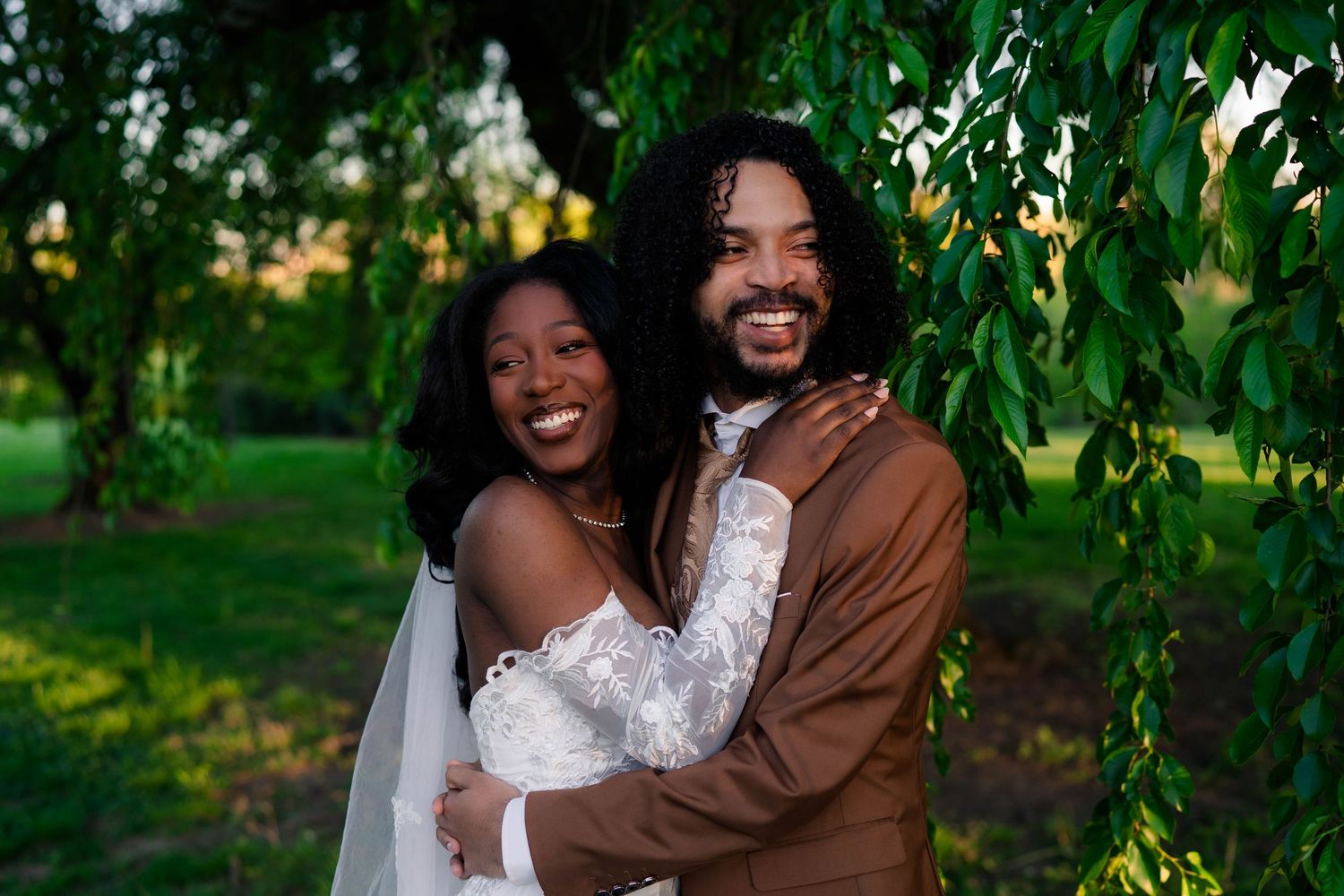 Joyful Black bride and groom embracing under weeping willow branches in a sunlit green garden.