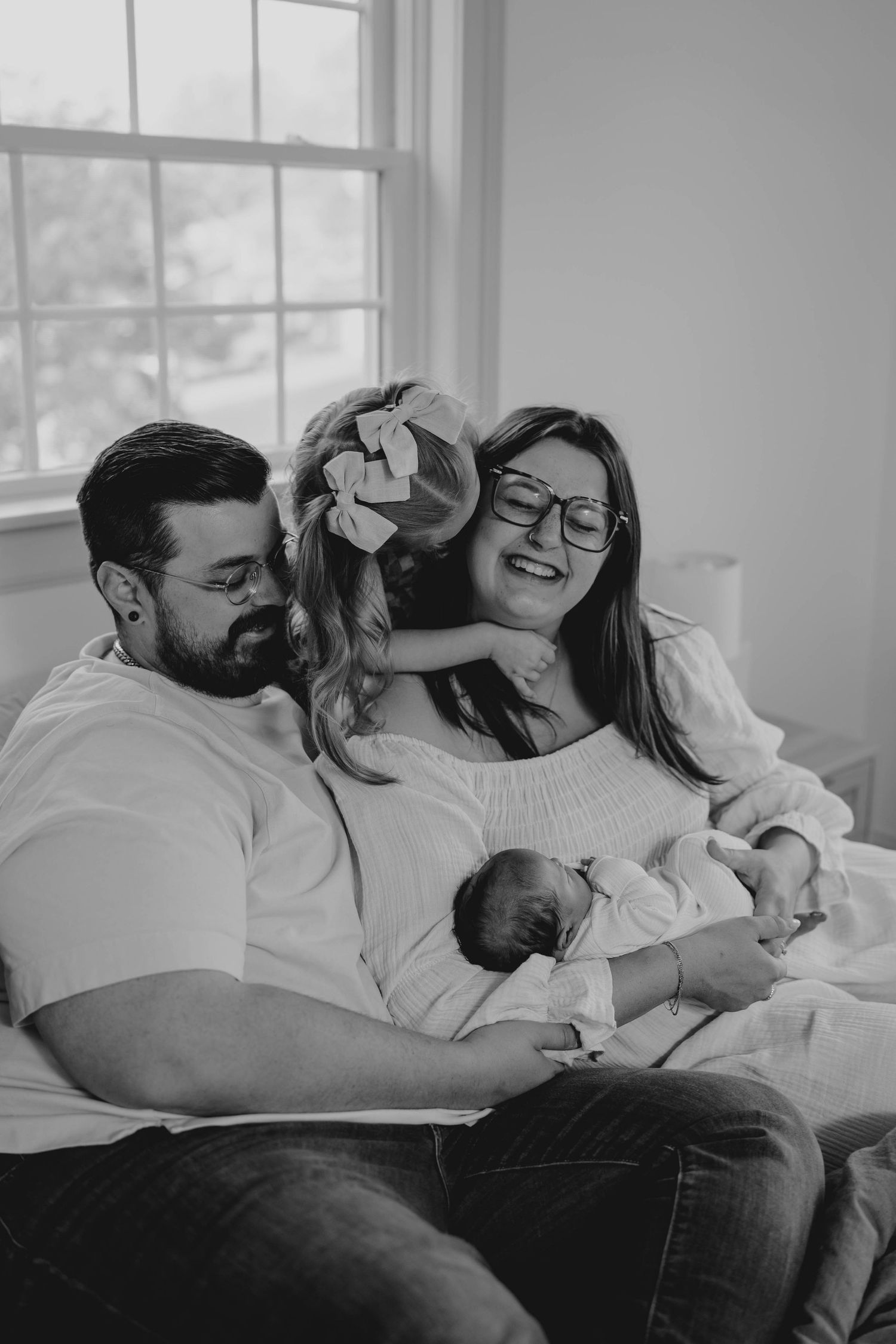 Black and white newborn family photo with parents and toddler daughter sitting together holding sleeping baby.