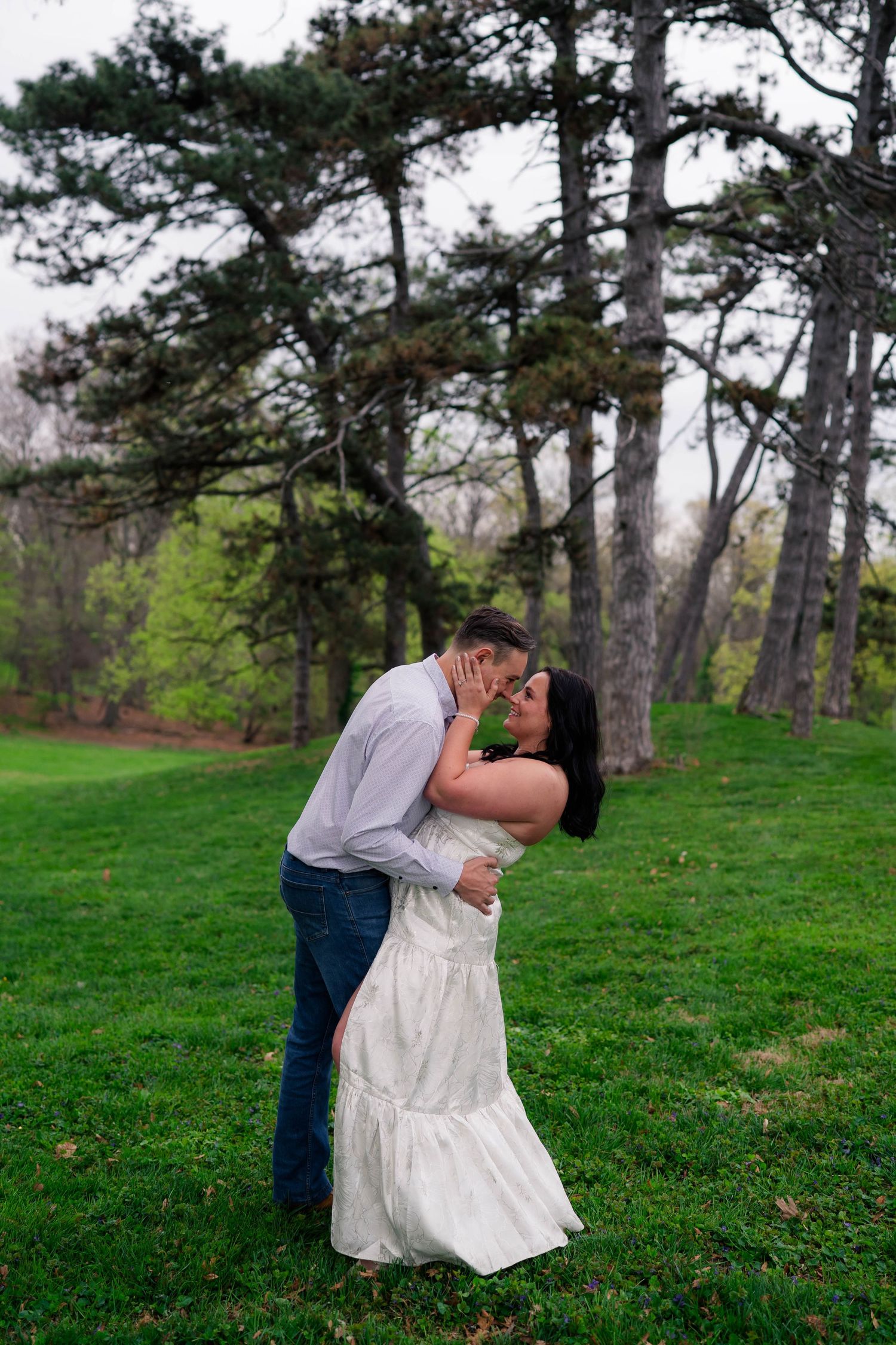 Couple sharing a romantic kiss on lush green grass with tall pine trees in the background.