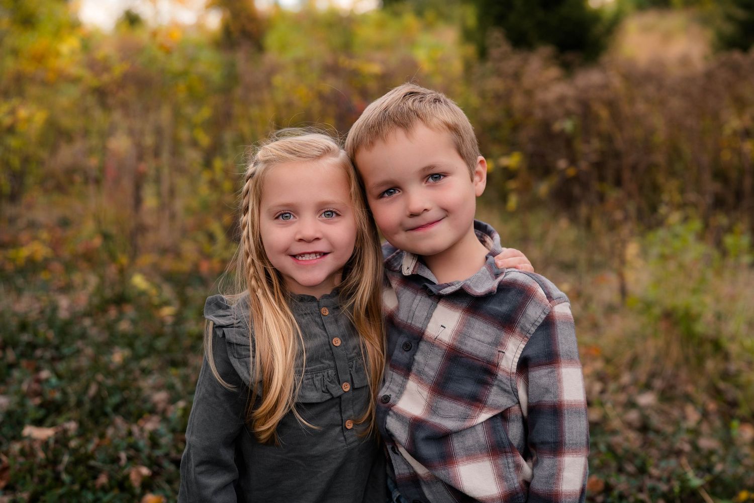 Two young siblings smiling together in a fall outdoor setting with colorful foliage background.