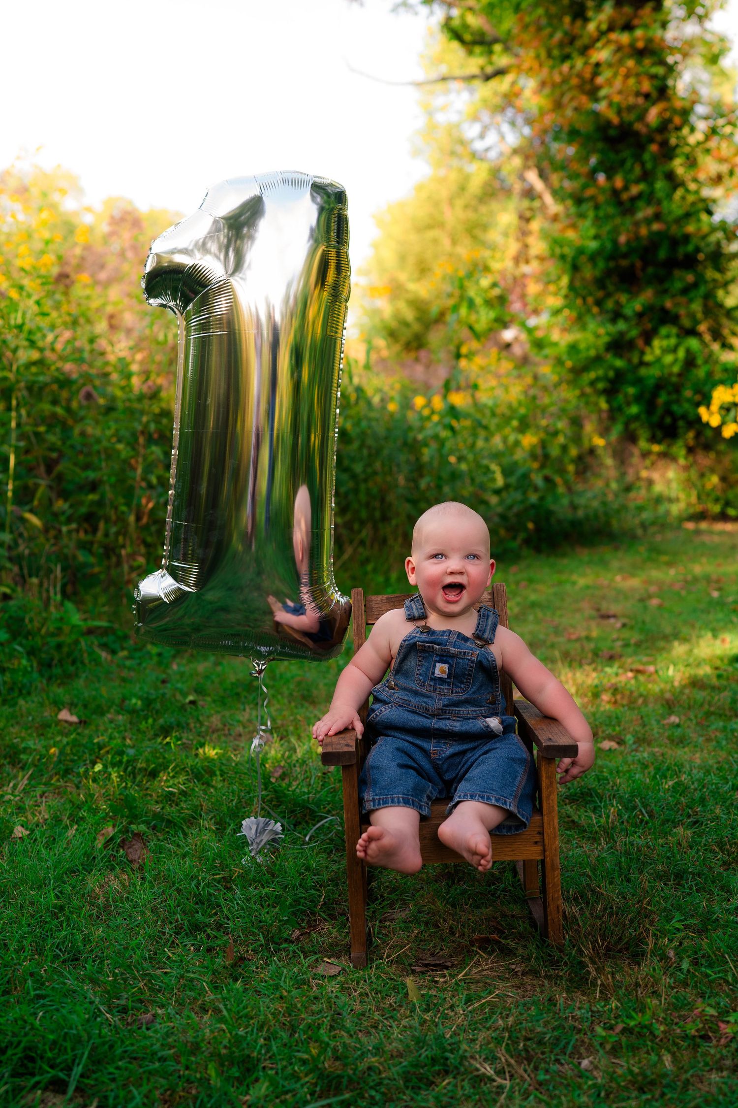 Laughing baby boy in denim overalls celebrating first birthday with a silver number one balloon outdoors.