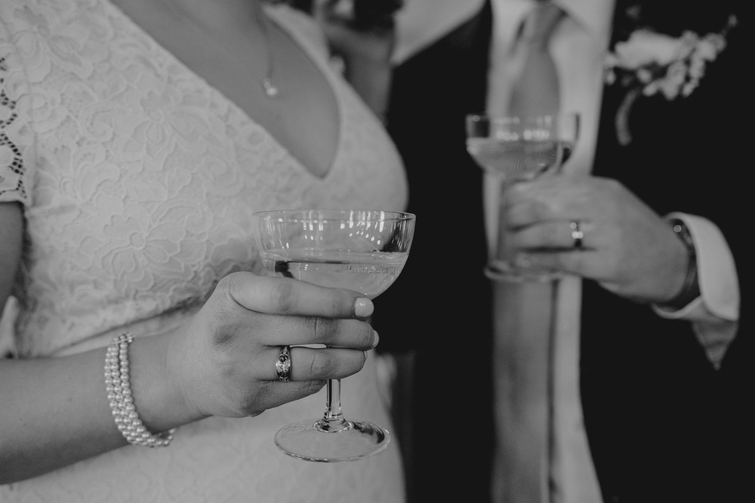 Black and white close-up of bride and groom holding champagne coupe glasses at wedding reception.