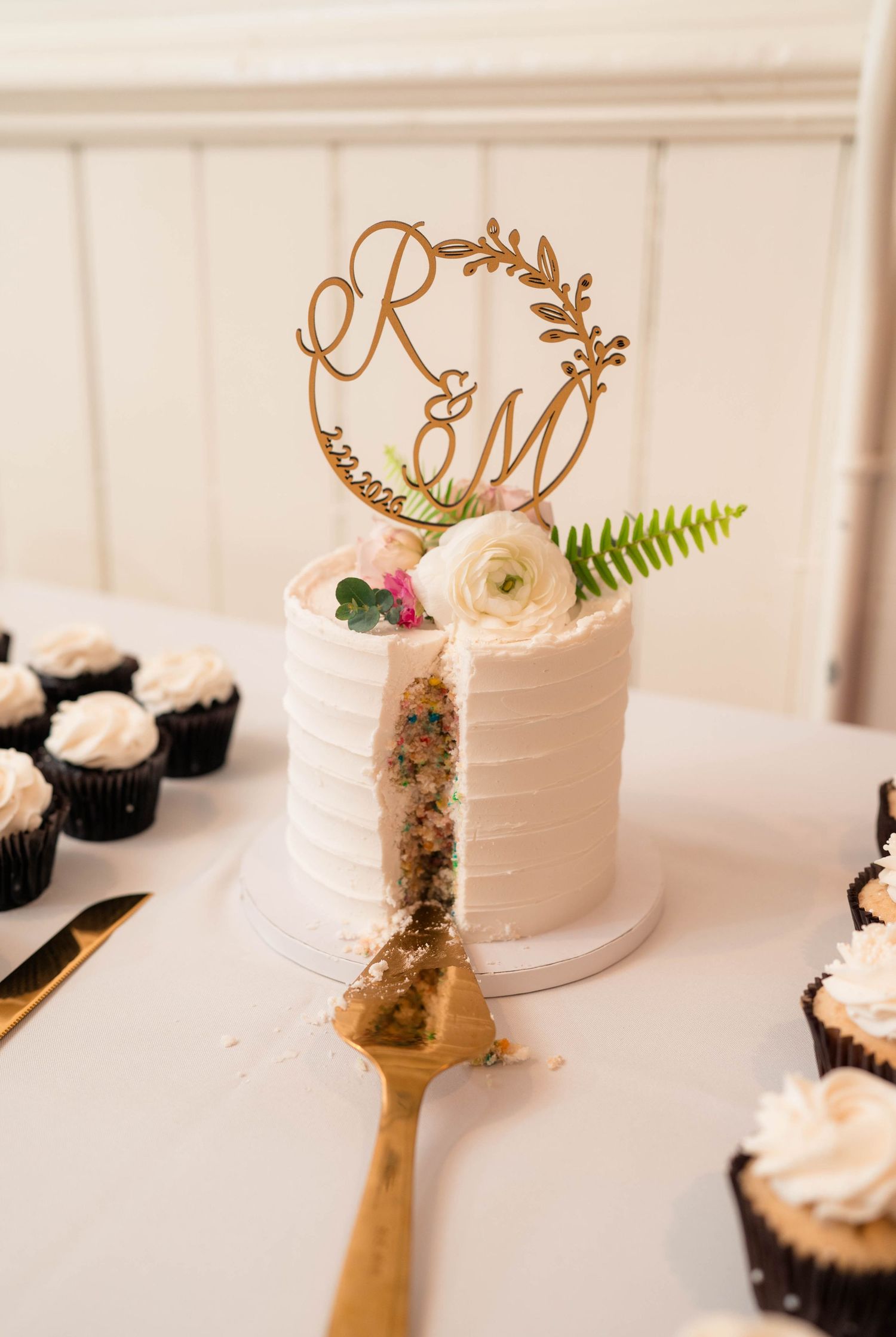 Sliced white wedding cake with confetti interior revealing colorful funfetti layers and wooden monogram topper.