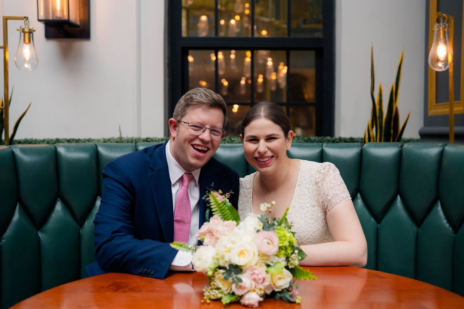 Joyful bride and groom smile together at table with pink and white floral wedding bouquet.