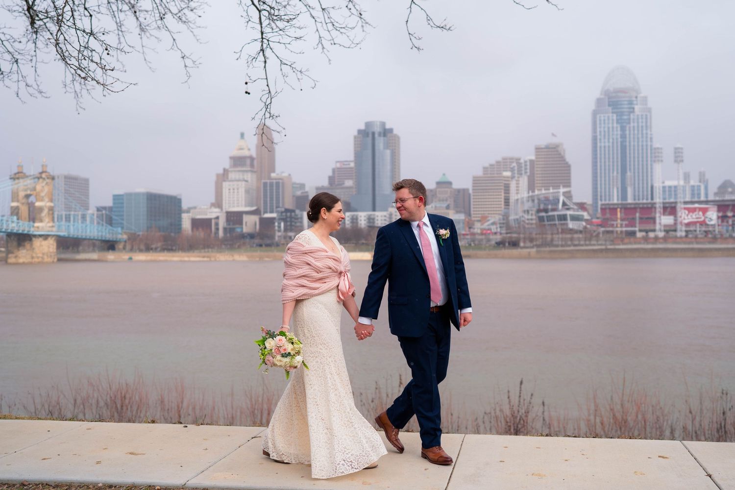 Wedding couple walking hand-in-hand along riverfront with Cincinnati skyline in misty background.