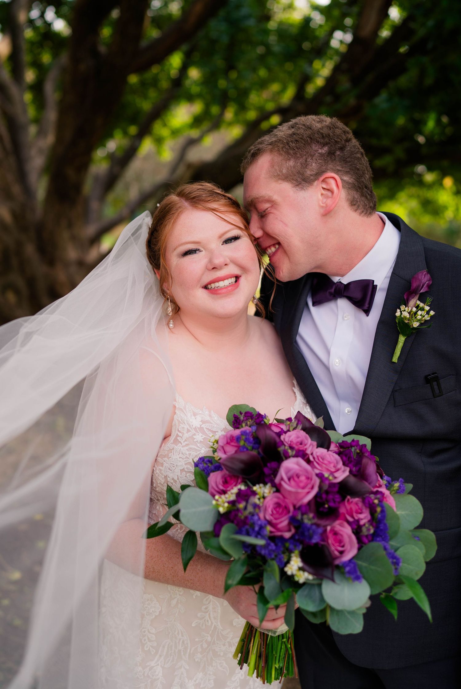 Happy bride and groom pose together outdoors, bride holding purple and pink rose bouquet.