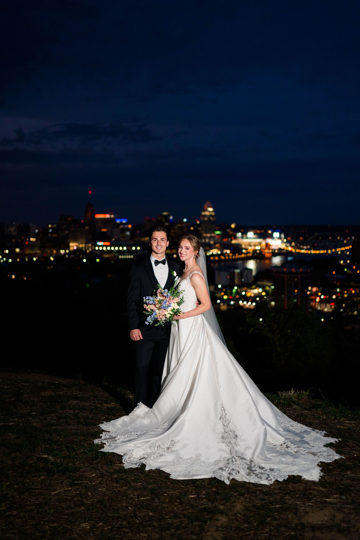 Bride and groom posing romantically with a glowing city skyline backdrop during their evening wedding portraits.