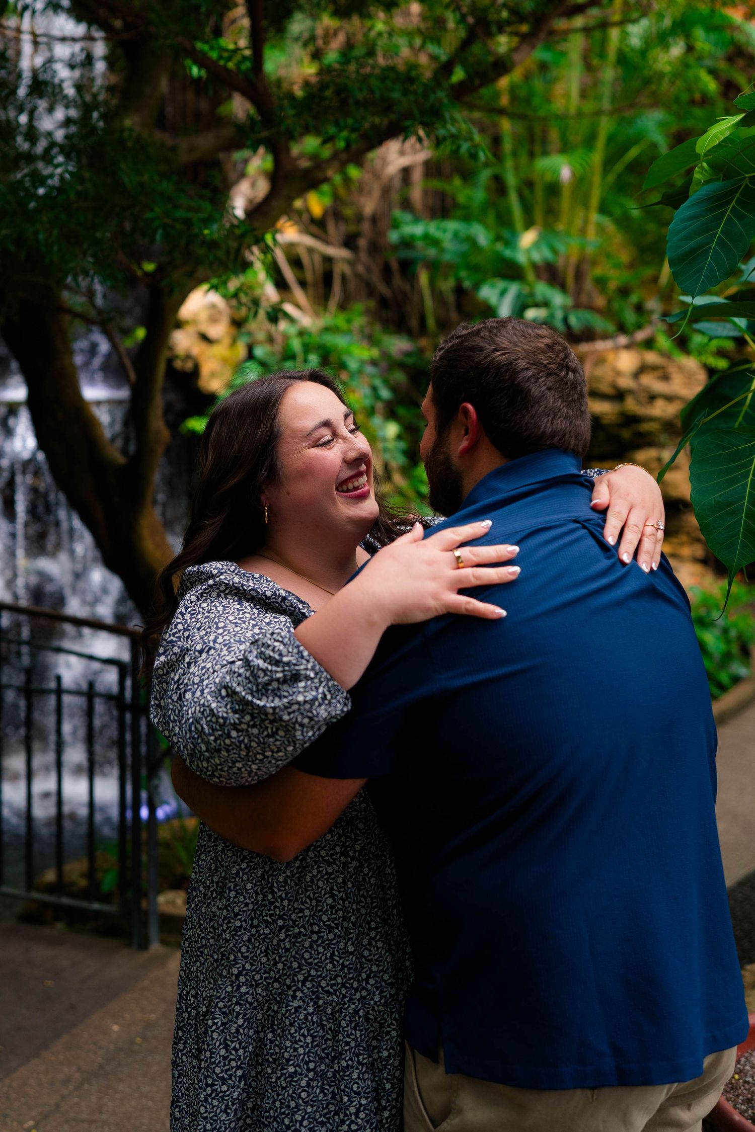 Joyful couple embracing near a waterfall in a lush green garden, woman laughing in a floral dress.