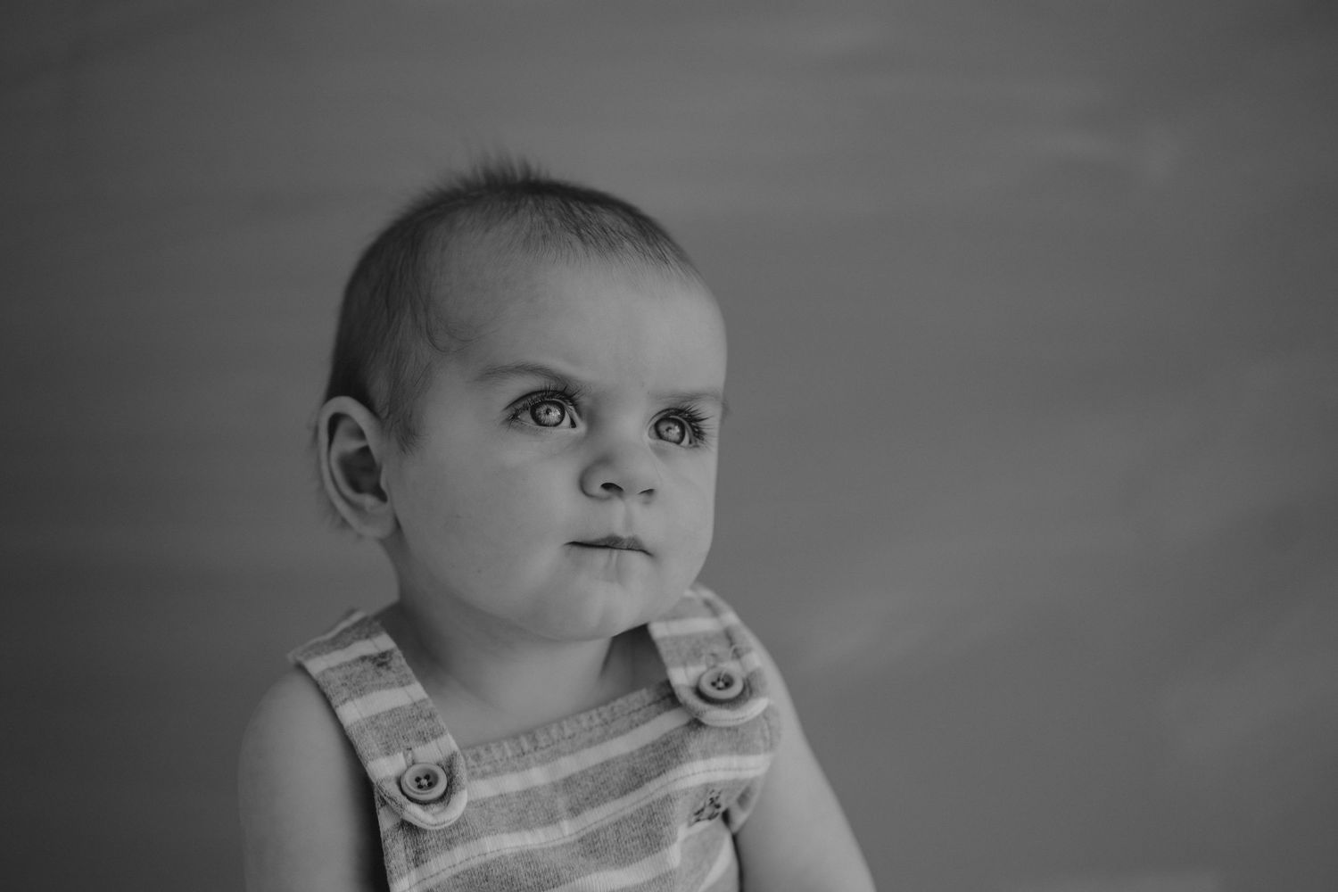 Black and white portrait of baby in striped overalls looking up thoughtfully.