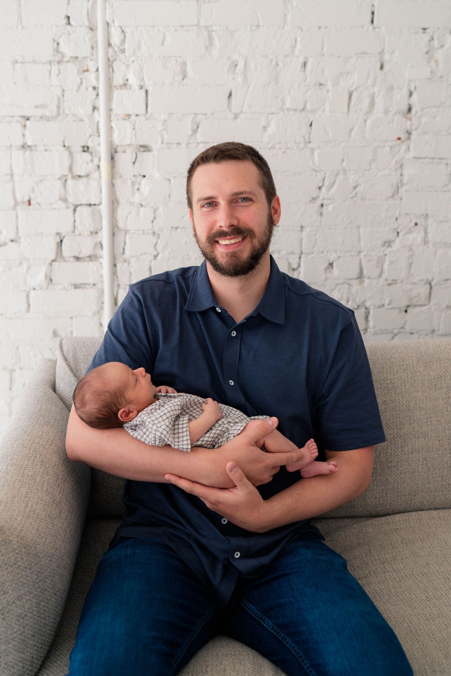 Smiling bearded father holding sleeping newborn baby while sitting on a couch against a white brick wall.