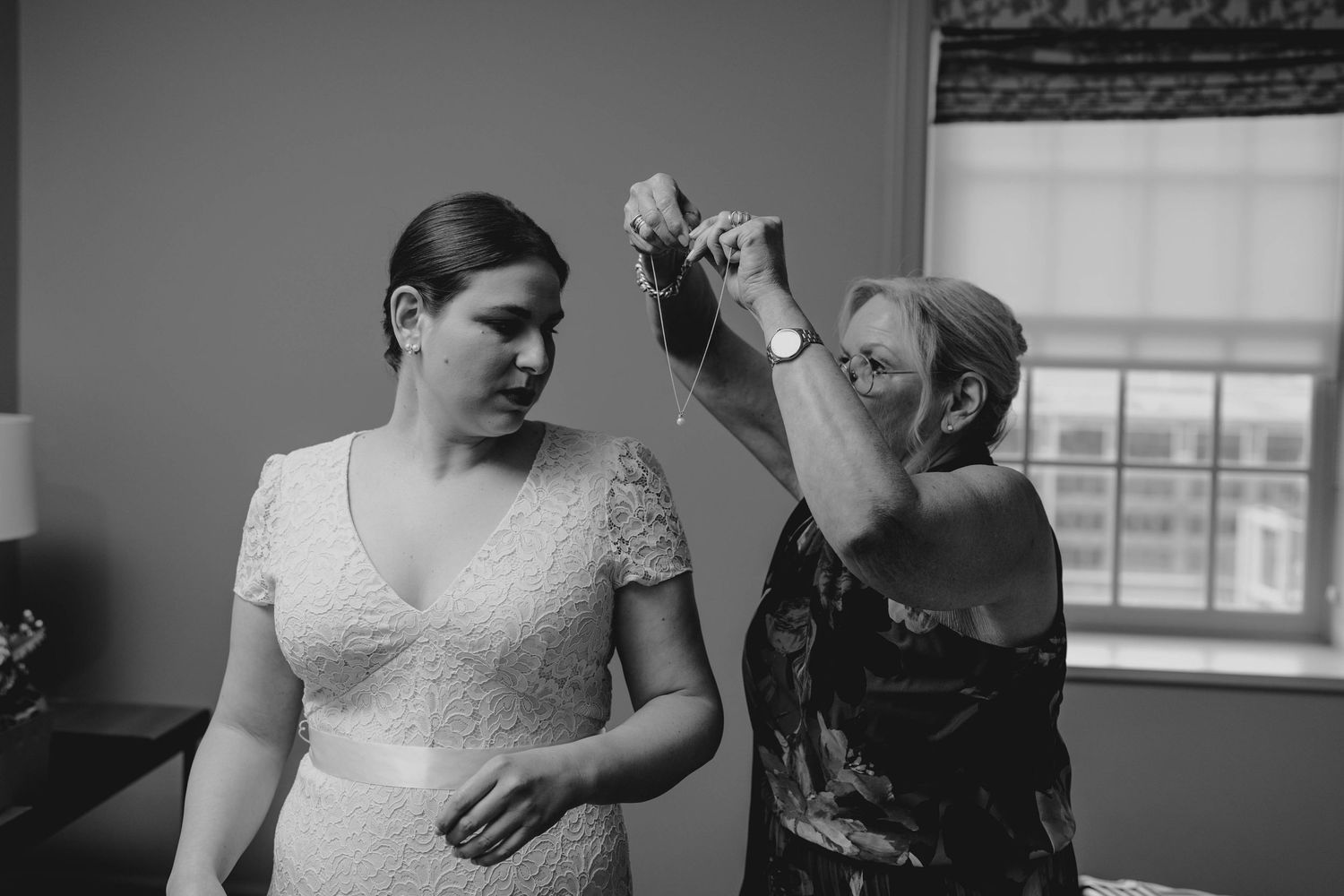Black and white photo of mother helping bride put on necklace before wedding.