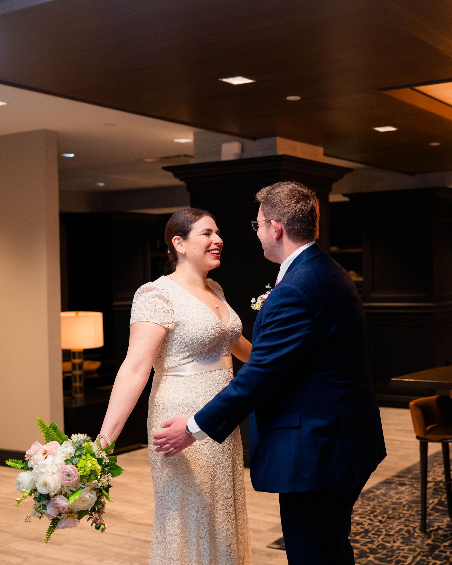Bride and groom laugh together during an intimate first look moment in a warmly lit hotel lobby.