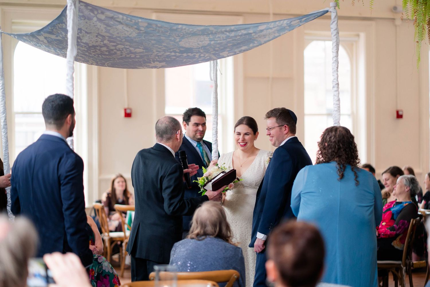 Wedding party gathers under blue chuppah as officiant reads during bright indoor ceremony.