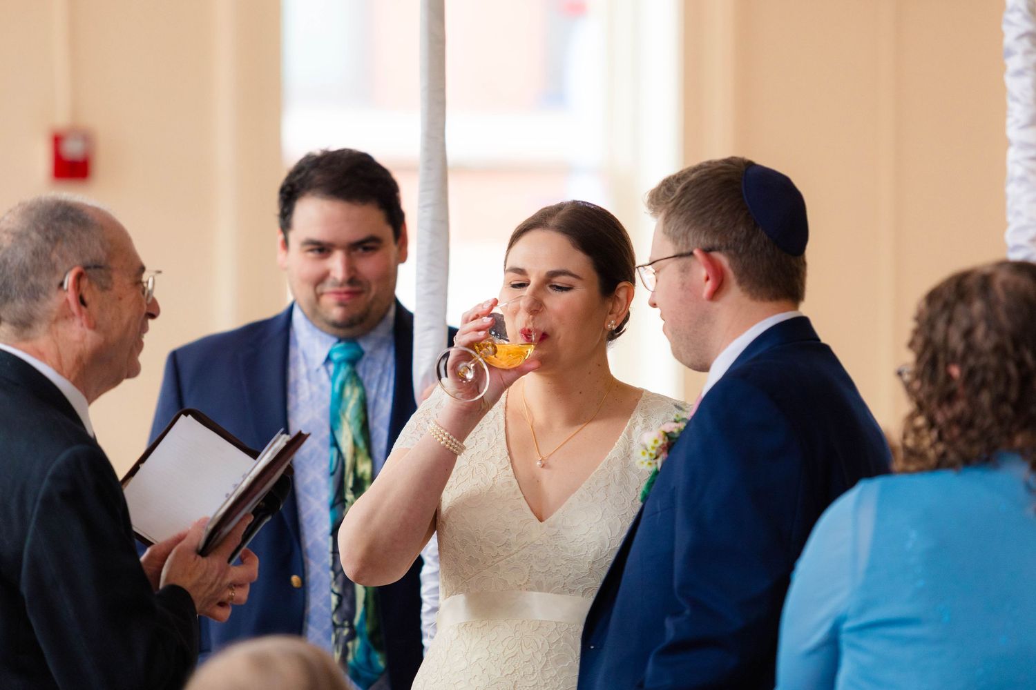Bride drinks from ceremonial cup during Jewish wedding ceremony while officiant holds book.