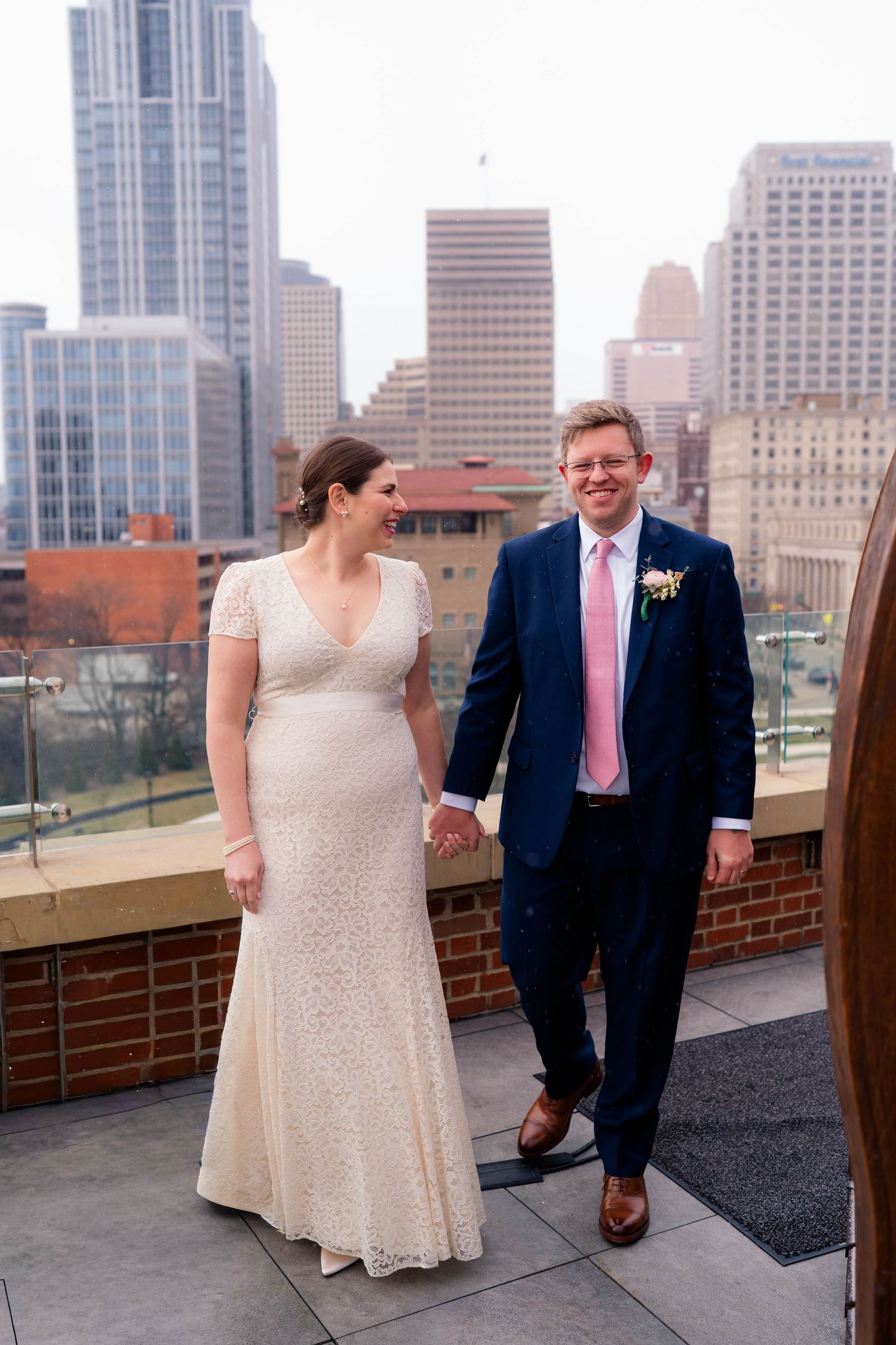 Bride and groom walking hand in hand on rooftop terrace with tall city buildings in background.