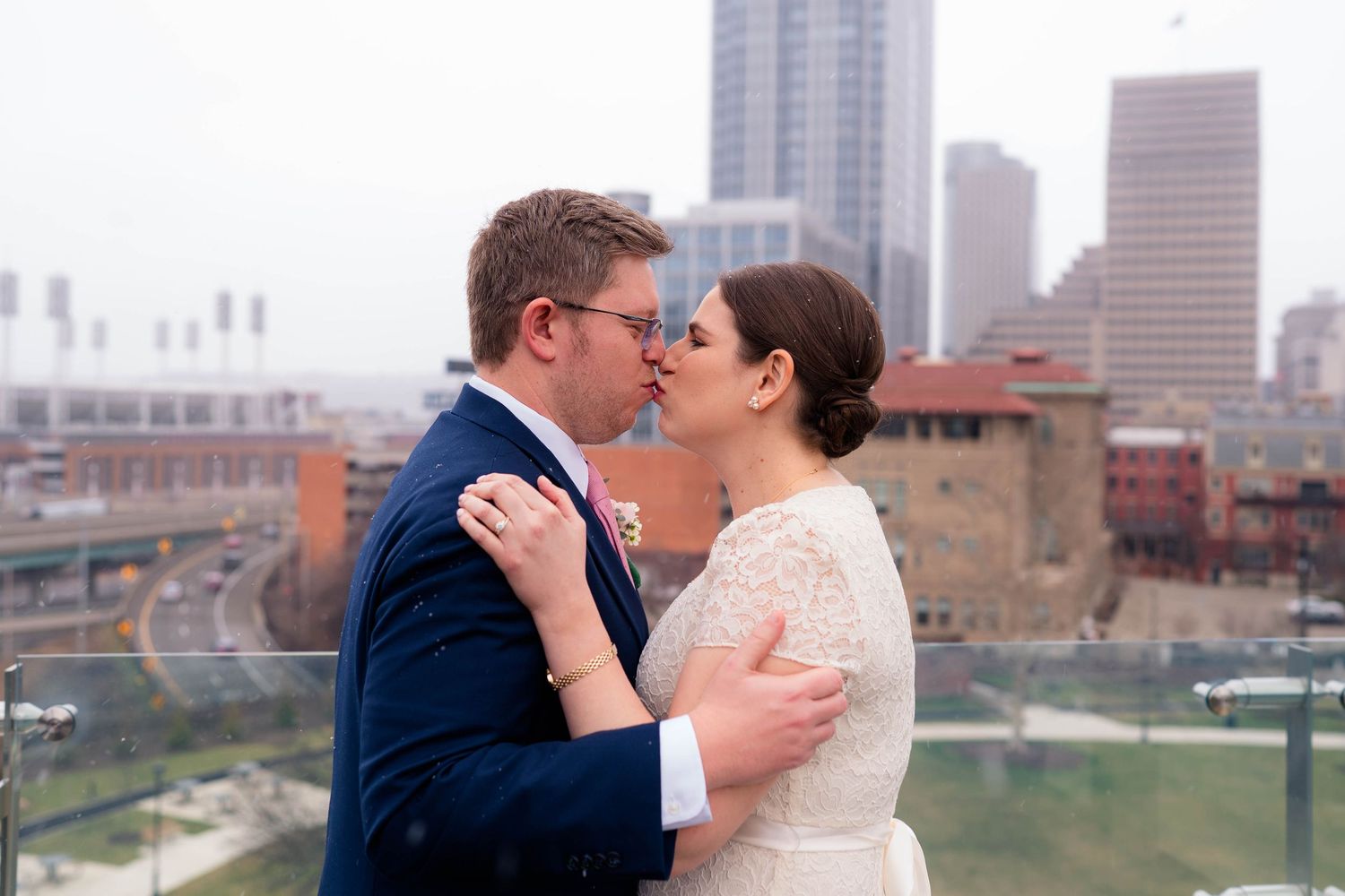 Bride and groom share a romantic rooftop kiss with a city skyline and baseball stadium visible behind them.