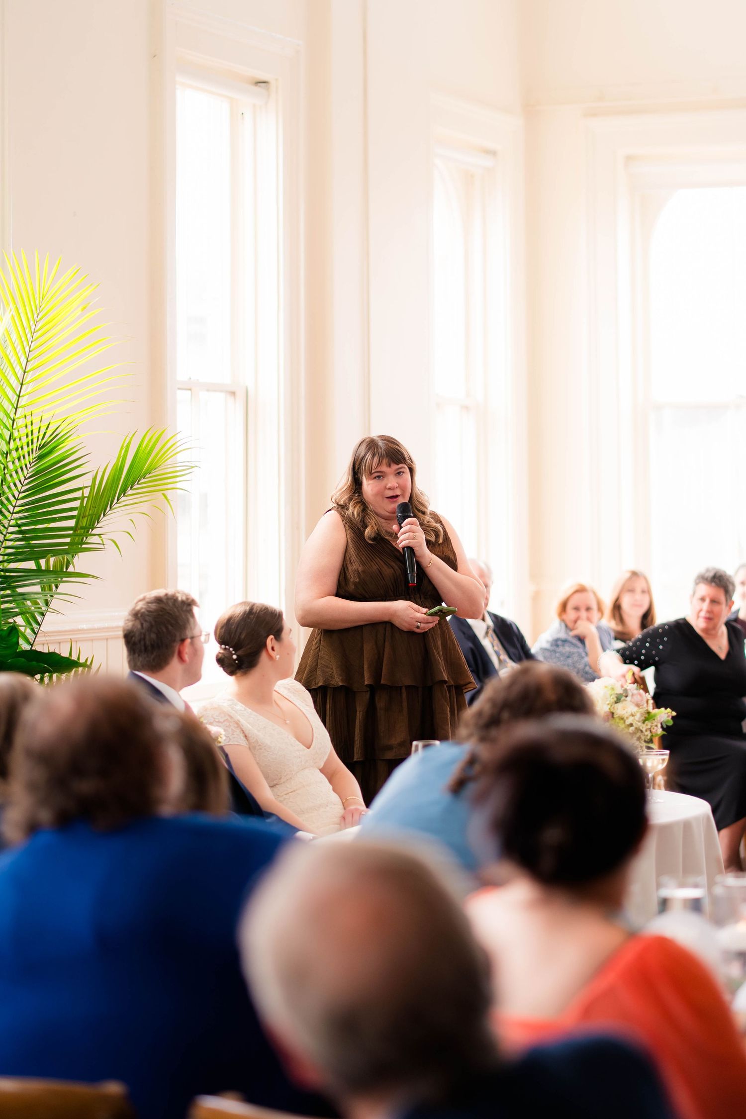 Woman in brown dress giving a wedding speech to seated guests in a bright elegant reception hall.