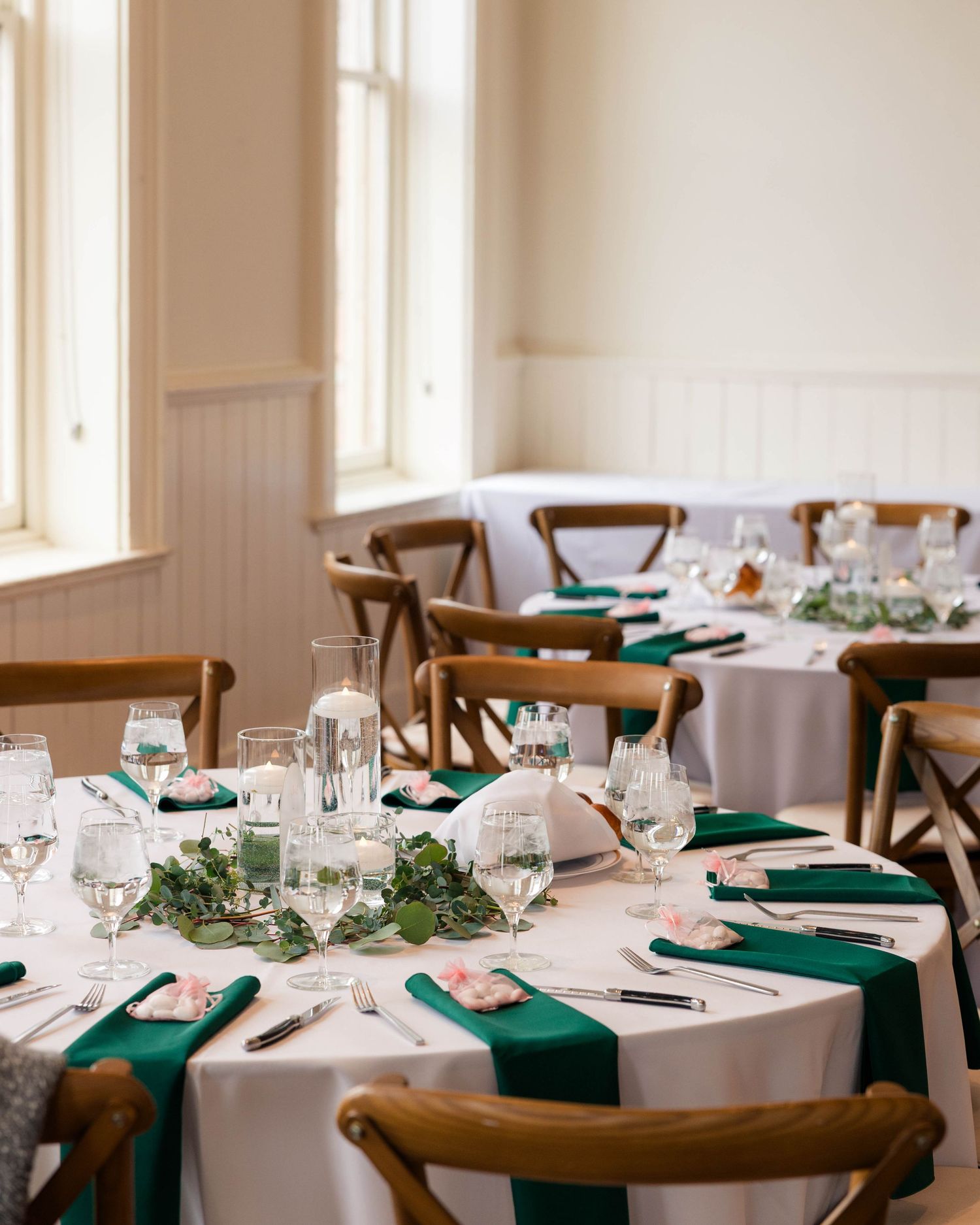 Elegant wedding reception tables set with emerald green napkins, glassware, and greenery centerpieces by bright windows.