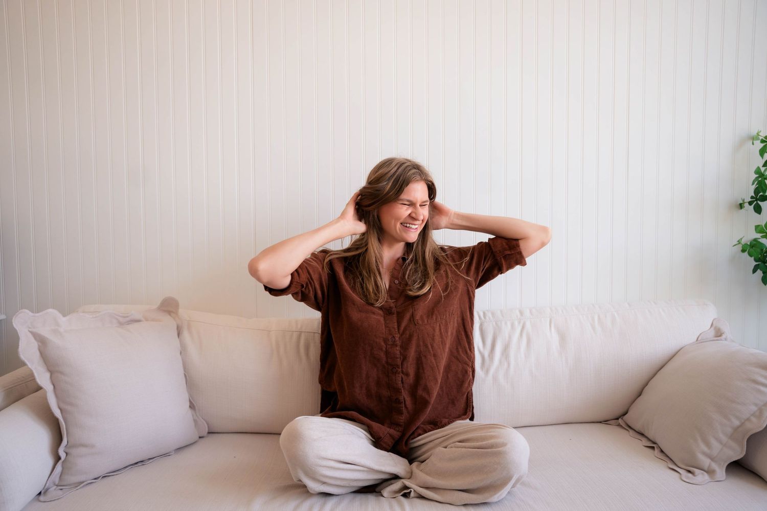 Happy woman sitting cross-legged on white couch with hands in hair.