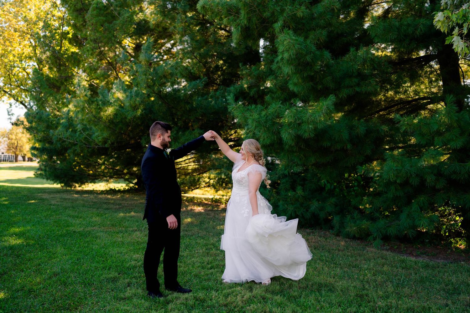 Groom twirls bride in flowing white gown on lush green grass beneath tall trees.